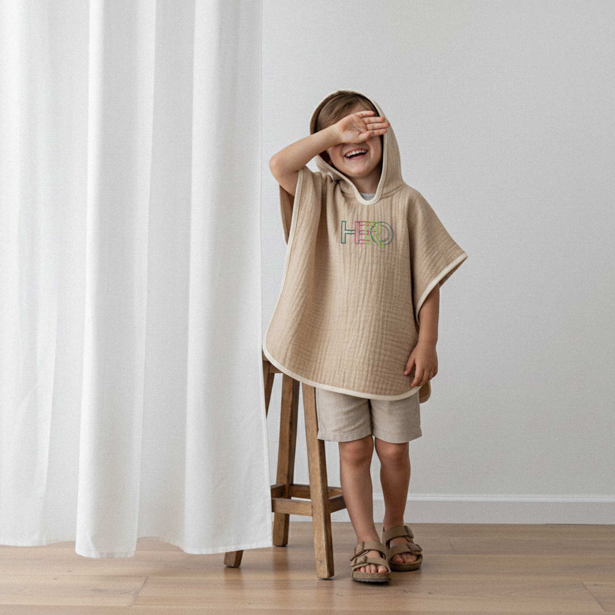 Bej şort ve somon rengi, 'HERO' nakışlı kapüşonlu müslin panço giyen erkek çocuk, bir gözünü kapatarak neşeyle kahkaha atıyor.