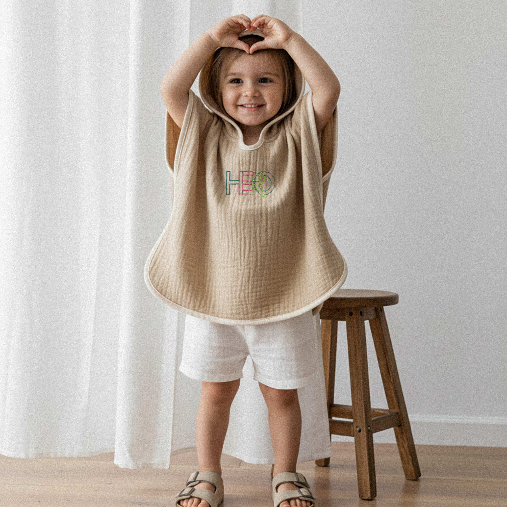 Beyaz şort ve somon rengi, 'HERO' nakışlı kapüşonlu müslin panço giyen kız çocuk, elleriyle kalp işareti yaparak gülümsüyor.