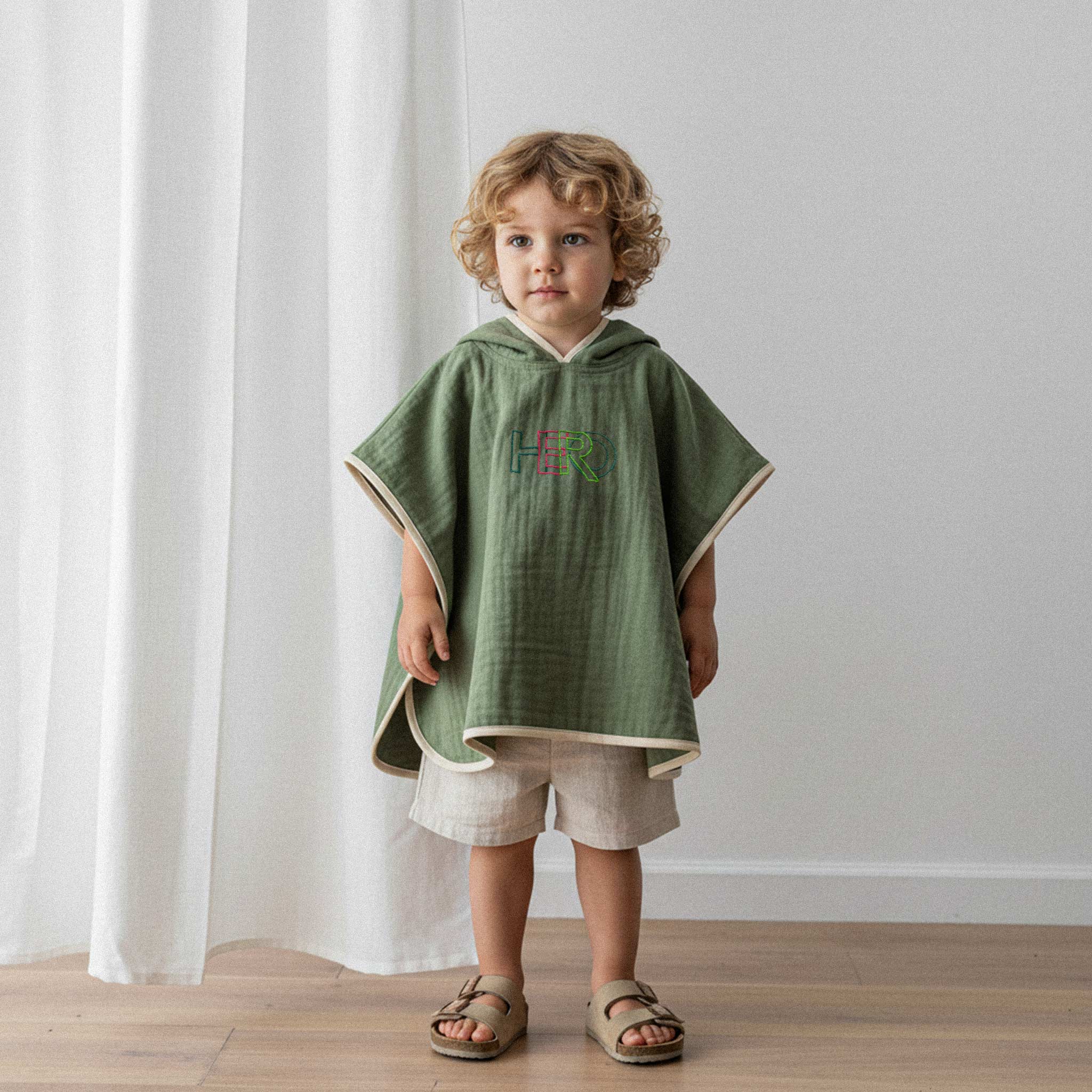 Kıvırcık saçlı küçük erkek çocuk, üzerinde haki yeşili, 'HERO' nakışlı kapüşonlu müslin panço ile sevimli bir şekilde poz veriyor.