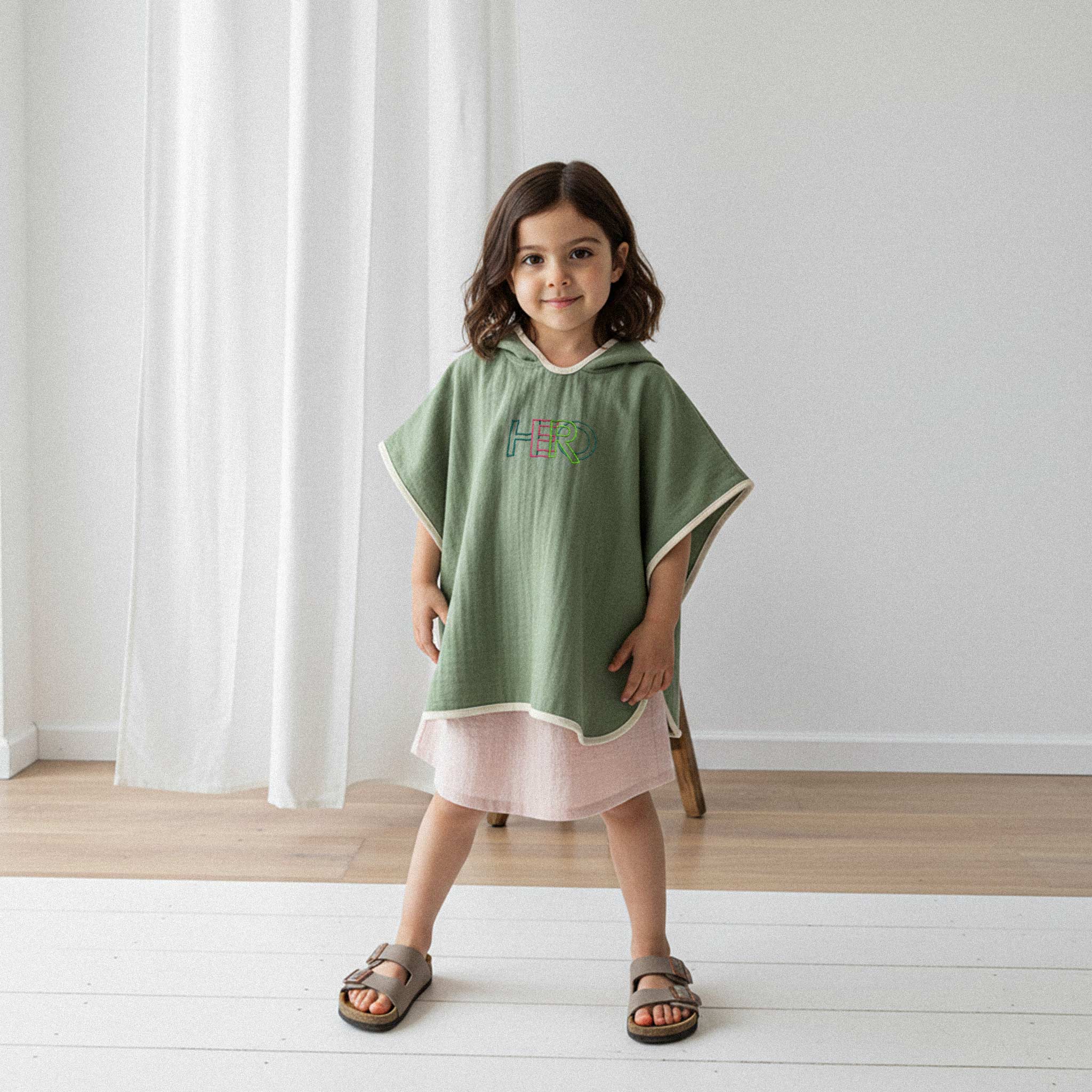 Pembe elbise üzerine haki yeşili, 'HERO' nakışlı kapüşonlu müslin panço giyen kız çocuk, gülümseyerek poz veriyor.