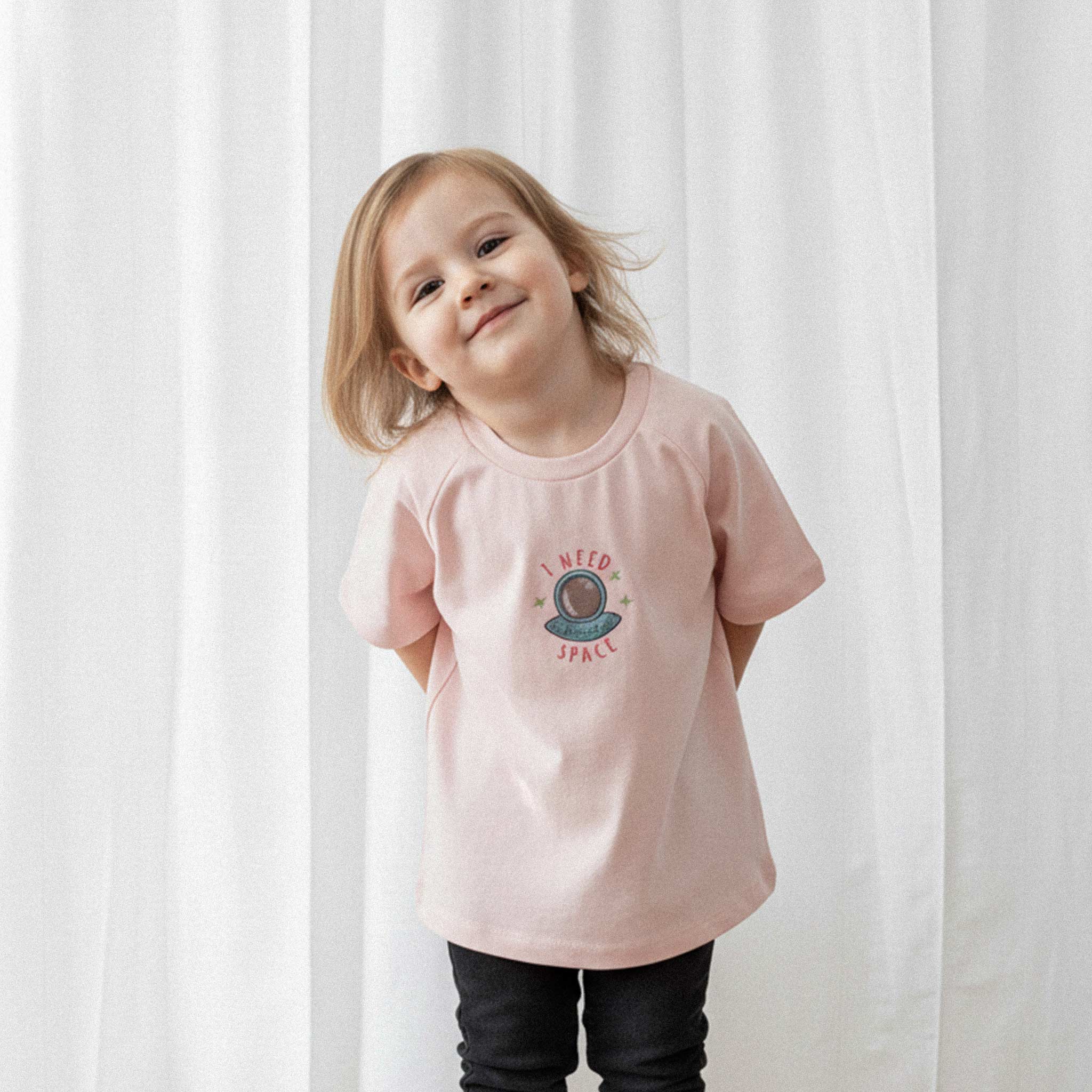 Siyah tayt ve gül kurusu, 'I Need Space' astronot nakışlı reglan kollu tişört giyen küçük kız çocuk, başını yana eğerek tatlı bir şekilde gülümsüyor.