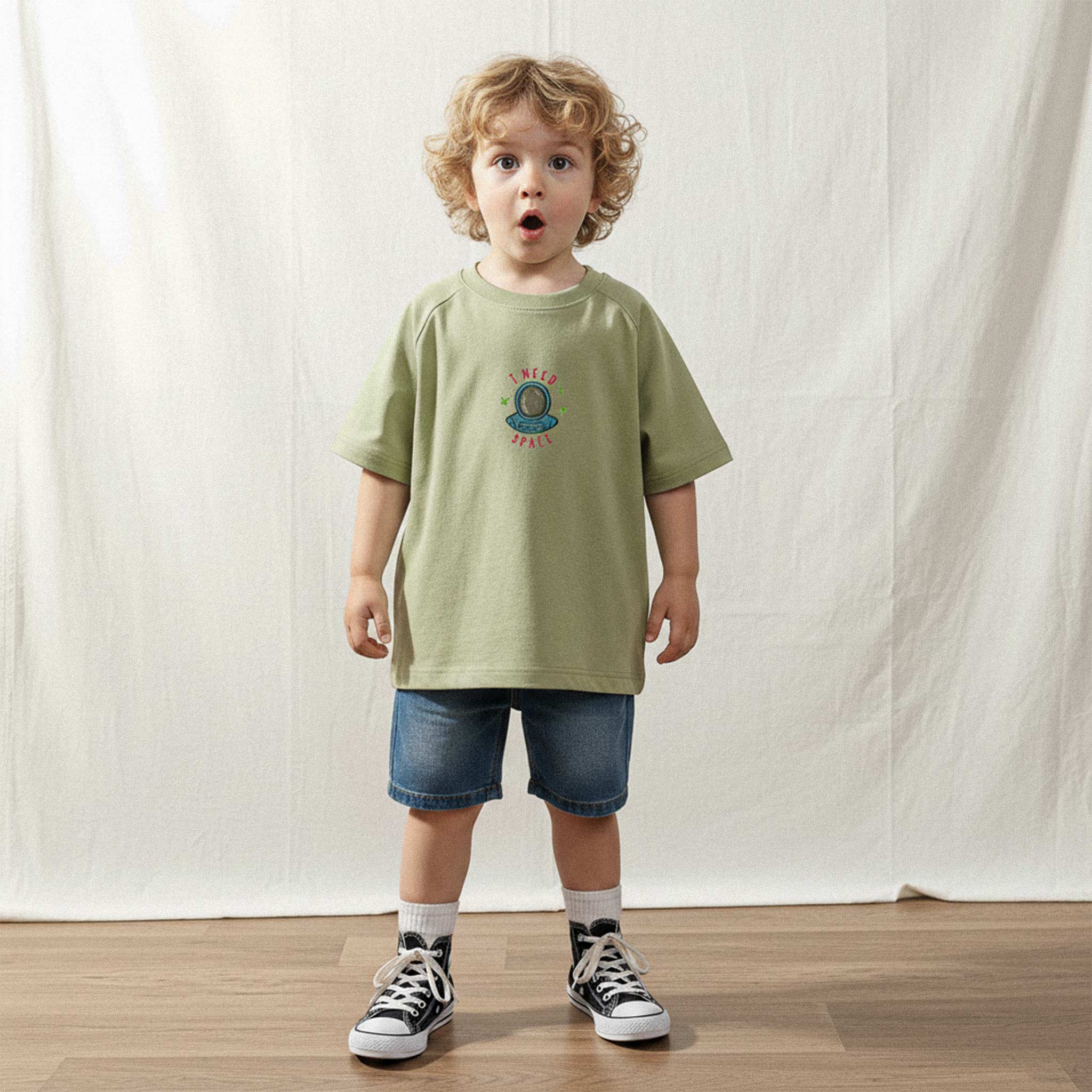 Üzerinde haki yeşili, reglan kollu, 'I Need Space' astronot nakışlı oversize bebek tişörtü bulunan kıvırcık saçlı küçük erkek çocuk, şaşkın ve sevimli bir ifadeyle kameraya bakıyor.
