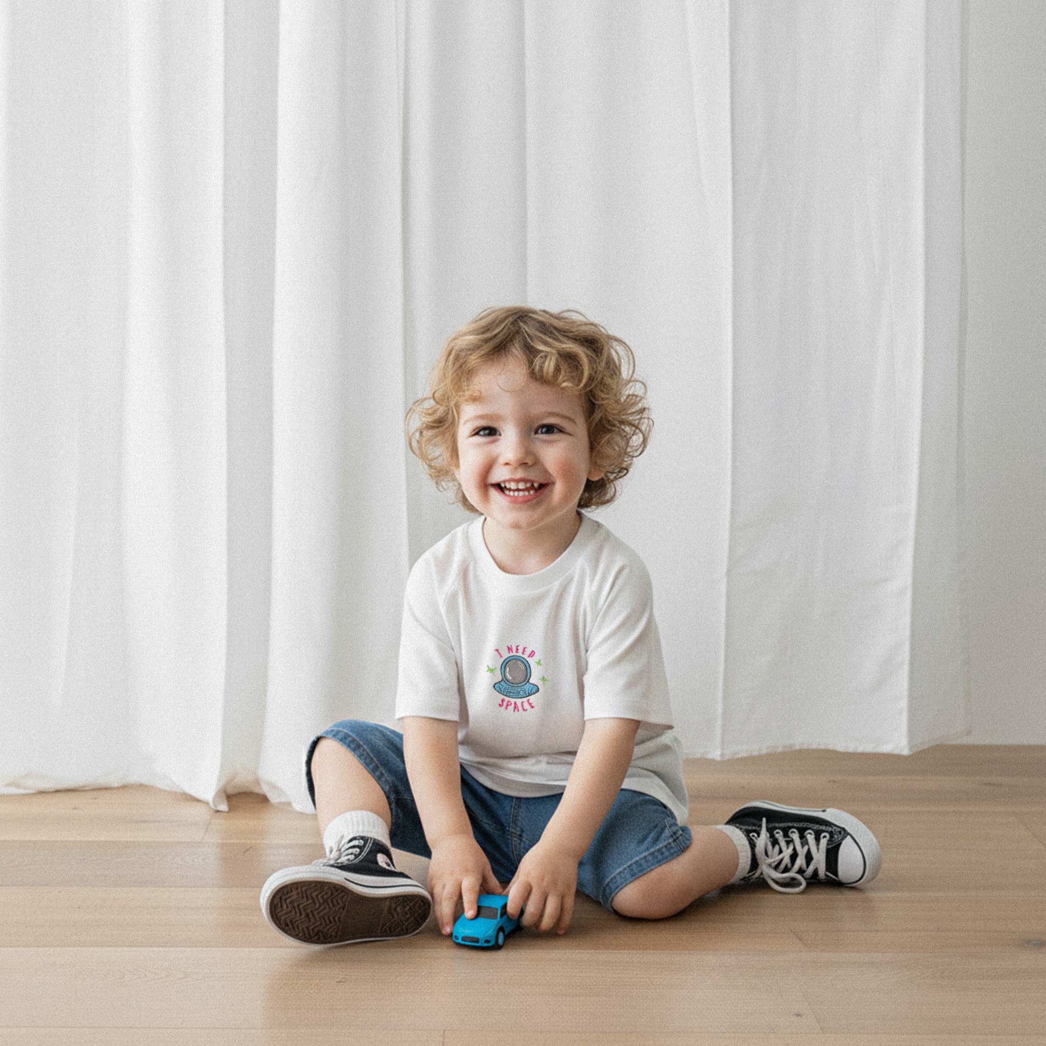 Jean şort ve ekru renkli, 'I Need Space' astronot nakışlı oversize tişört giyen kıvırcık saçlı küçük erkek çocuk, yerde oyuncak arabasıyla oynarken neşeyle gülümsüyor.