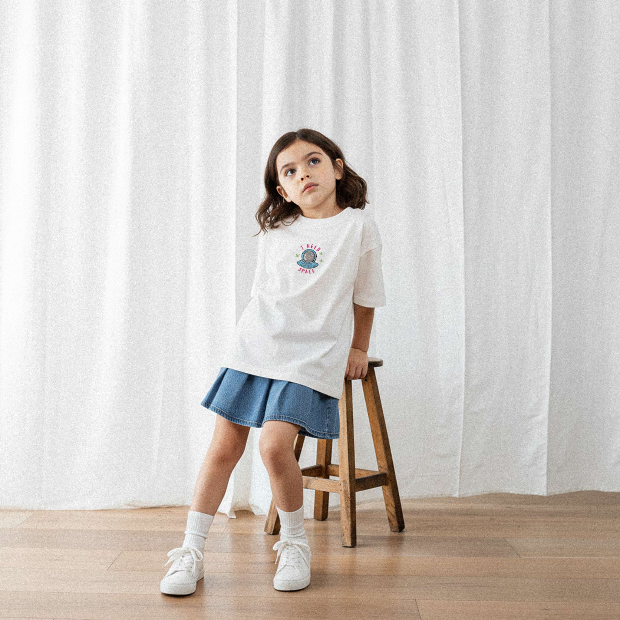 Jean etek ve ekru renkli, 'I Need Space' astronot nakışlı oversize tişört giyen kız çocuk, ahşap bir taburede otururken düşünceli bir şekilde yukarı bakıyor.