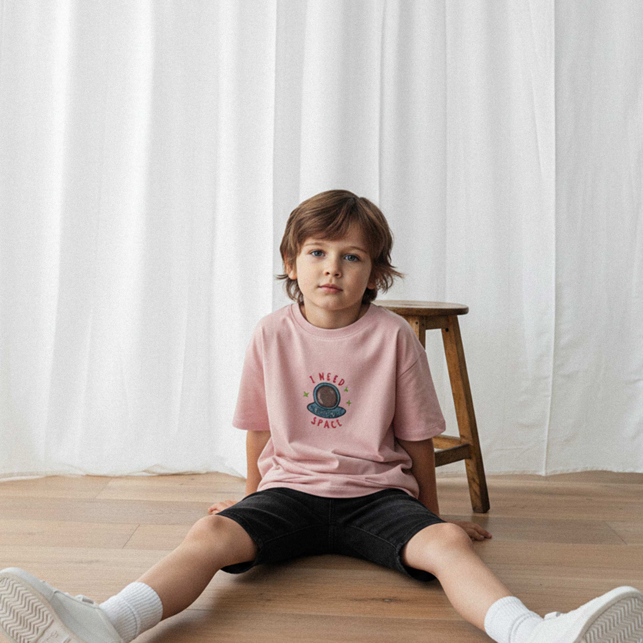 Siyah şort ve gül kurusu renkli, 'I Need Space' astronot nakışlı oversize tişört giyen erkek çocuk, yerde oturarak kameraya bakıyor.