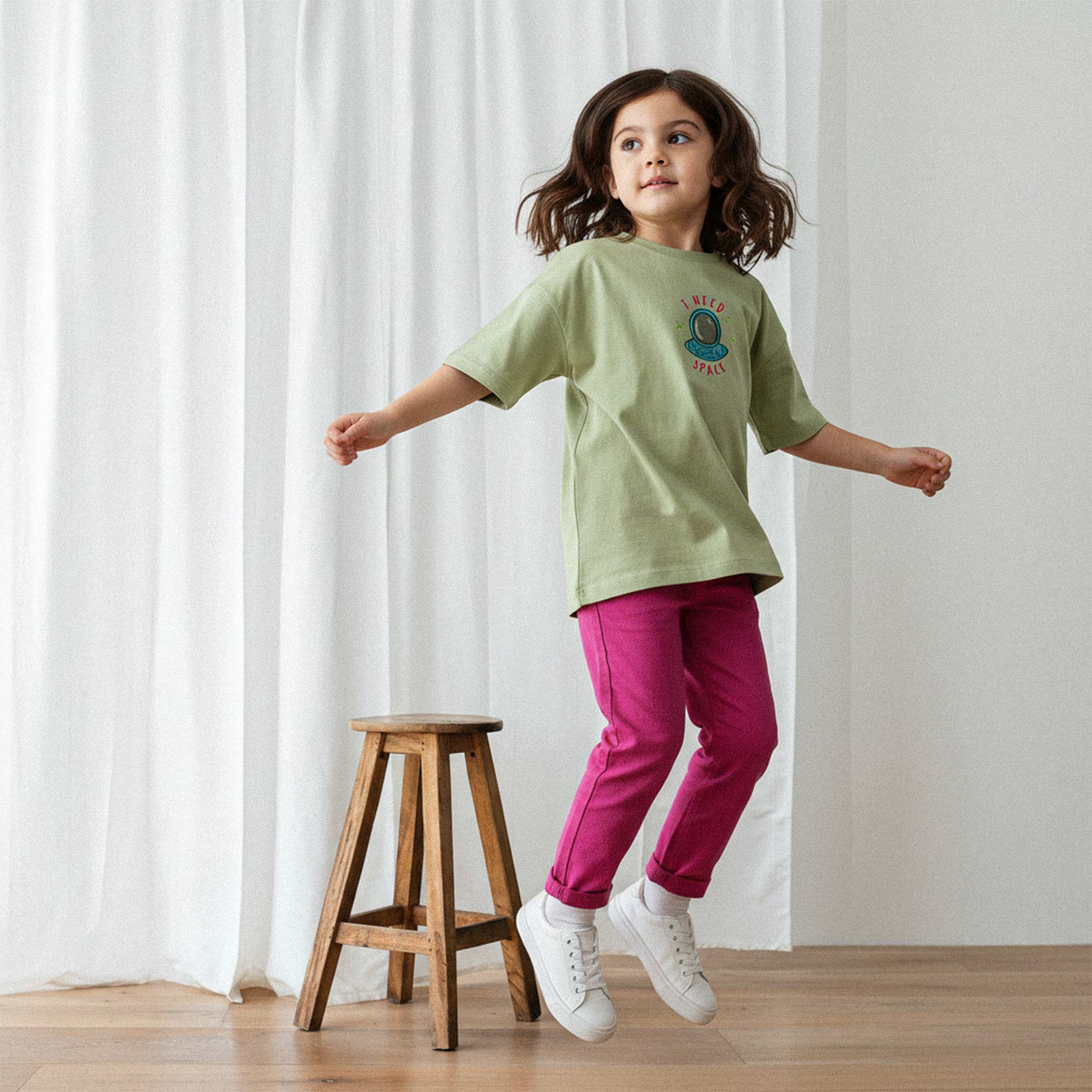Fuşya rengi tayt ve haki yeşili, 'I Need Space' astronot nakışlı oversize tişört giyen kız çocuk, enerjik bir şekilde zıplarken havada poz veriyor.