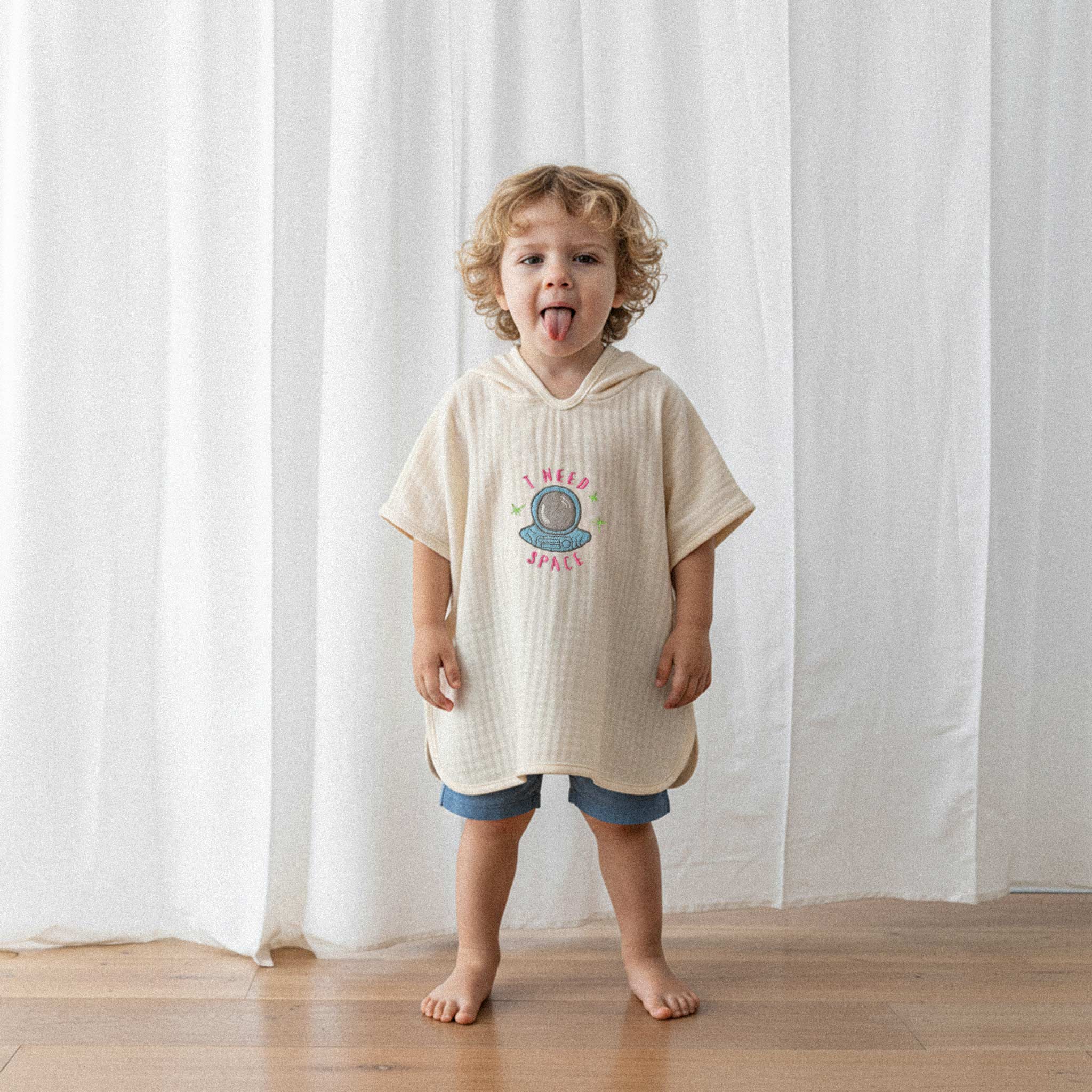 Üzerinde ekru renkli, 'I Need Space' astronot nakışlı kapüşonlu müslin panço bulunan kıvırcık saçlı küçük erkek çocuk, dilini çıkararak eğlenceli bir poz veriyor.