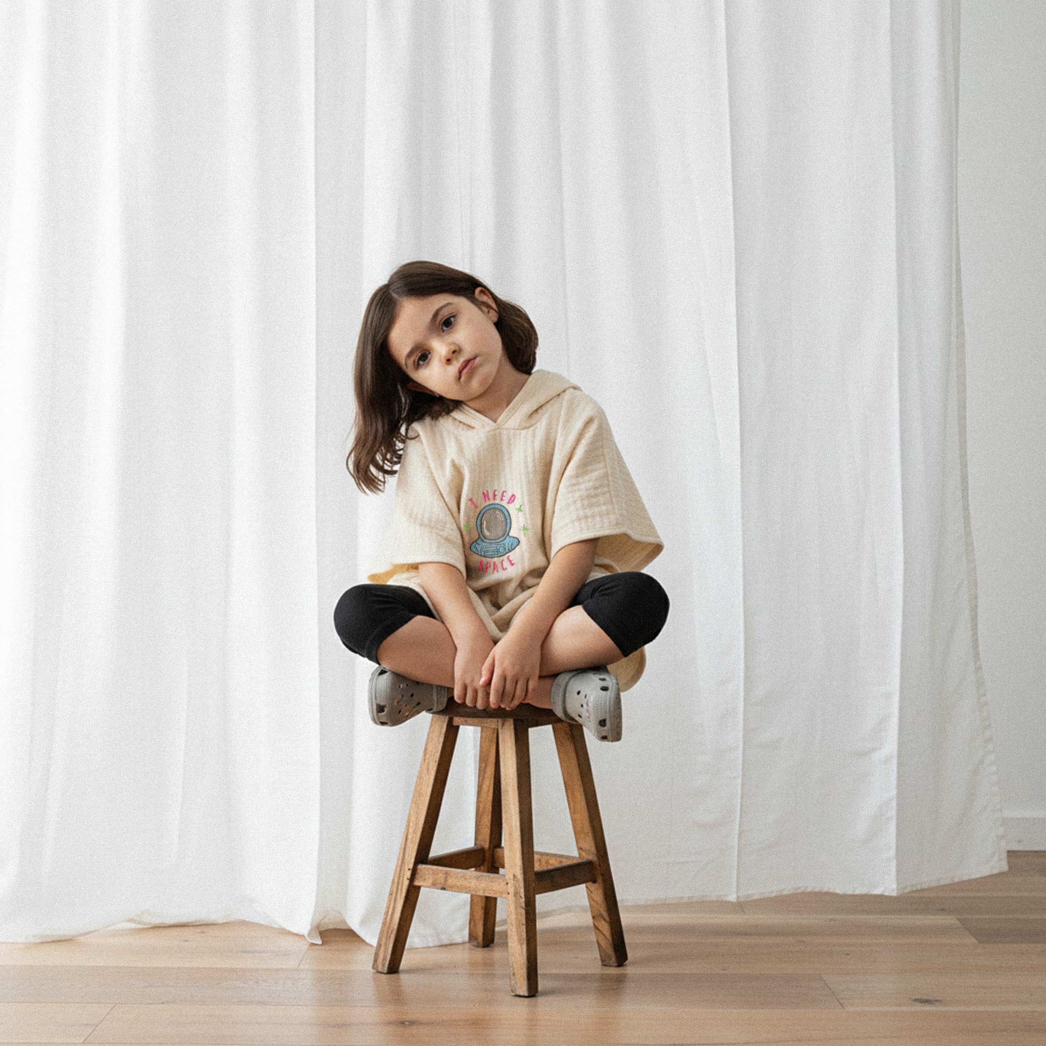 Siyah tayt ve ekru renkli, 'I Need Space' astronot nakışlı kapüşonlu müslin panço giyen kız çocuk, ahşap bir taburede oturarak havalı bir poz veriyor.