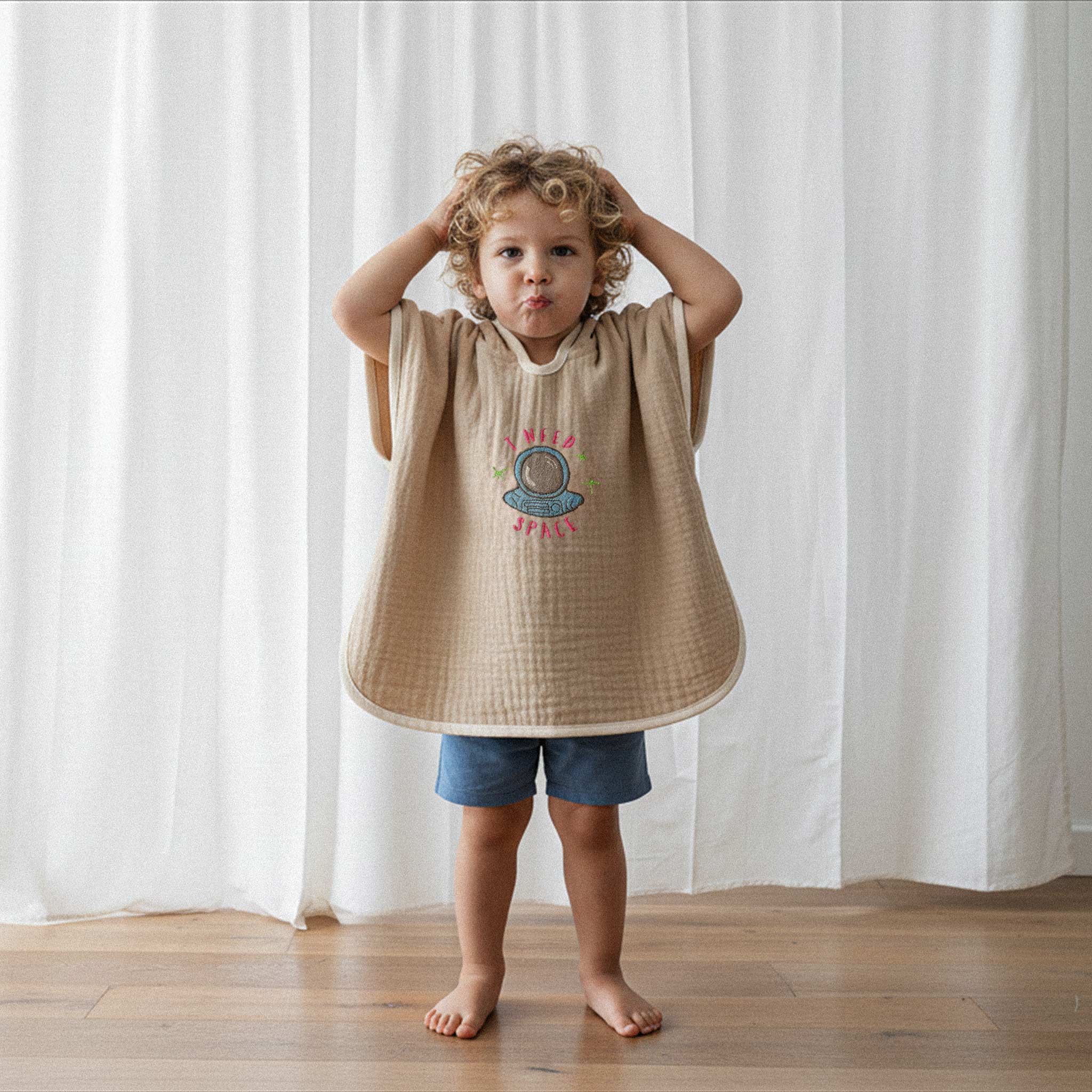 Mavi şort ve somon rengi, 'I Need Space' astronot nakışlı kapüşonlu müslin panço giyen kıvırcık saçlı küçük erkek çocuk, elleri başında komik bir surat ifadesi yapıyor.