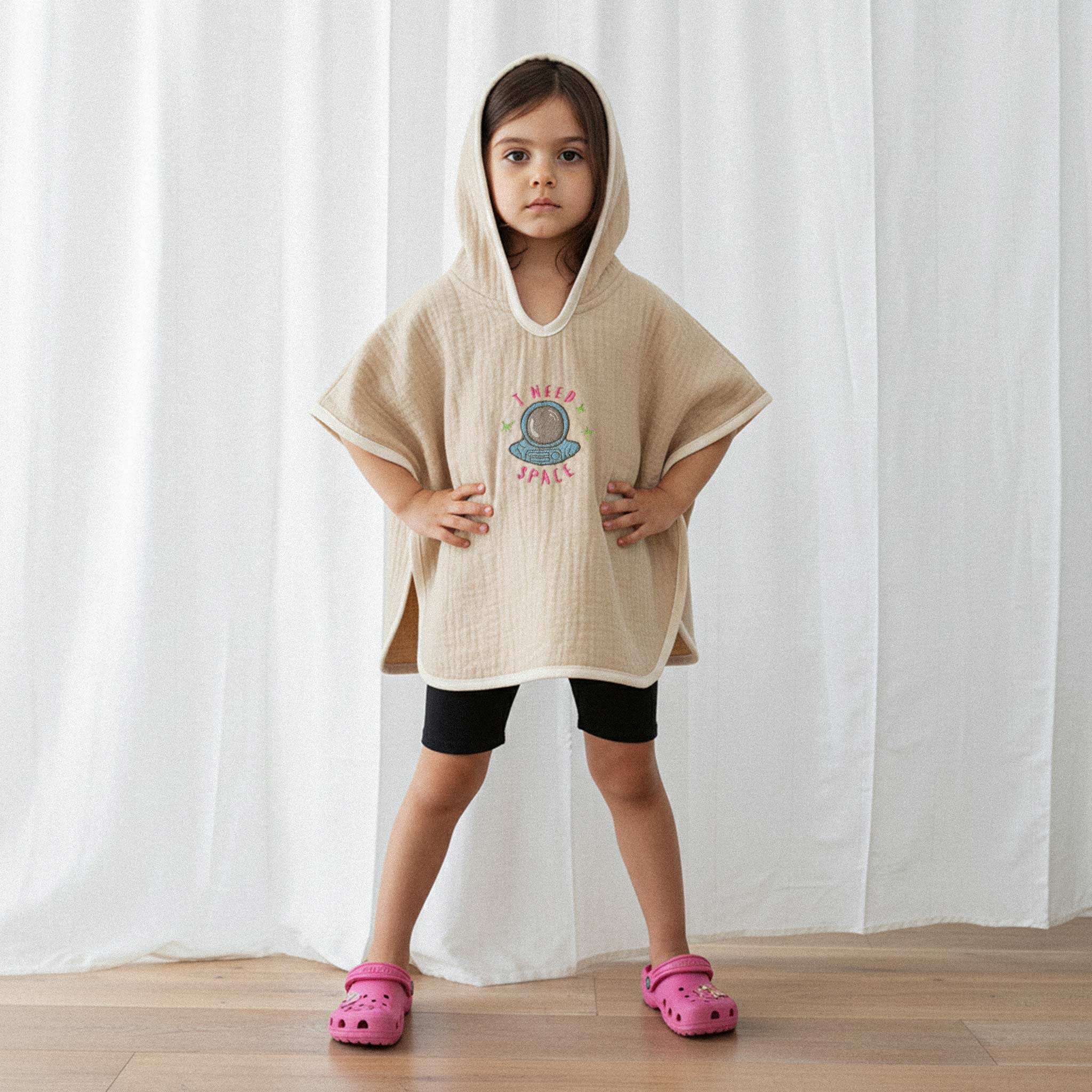 Siyah şort ve pembe terlik giyen kız çocuk, üzerinde somon rengi, 'I Need Space' astronot nakışlı kapüşonlu müslin panço ile elleri belinde havalı bir poz veriyor.