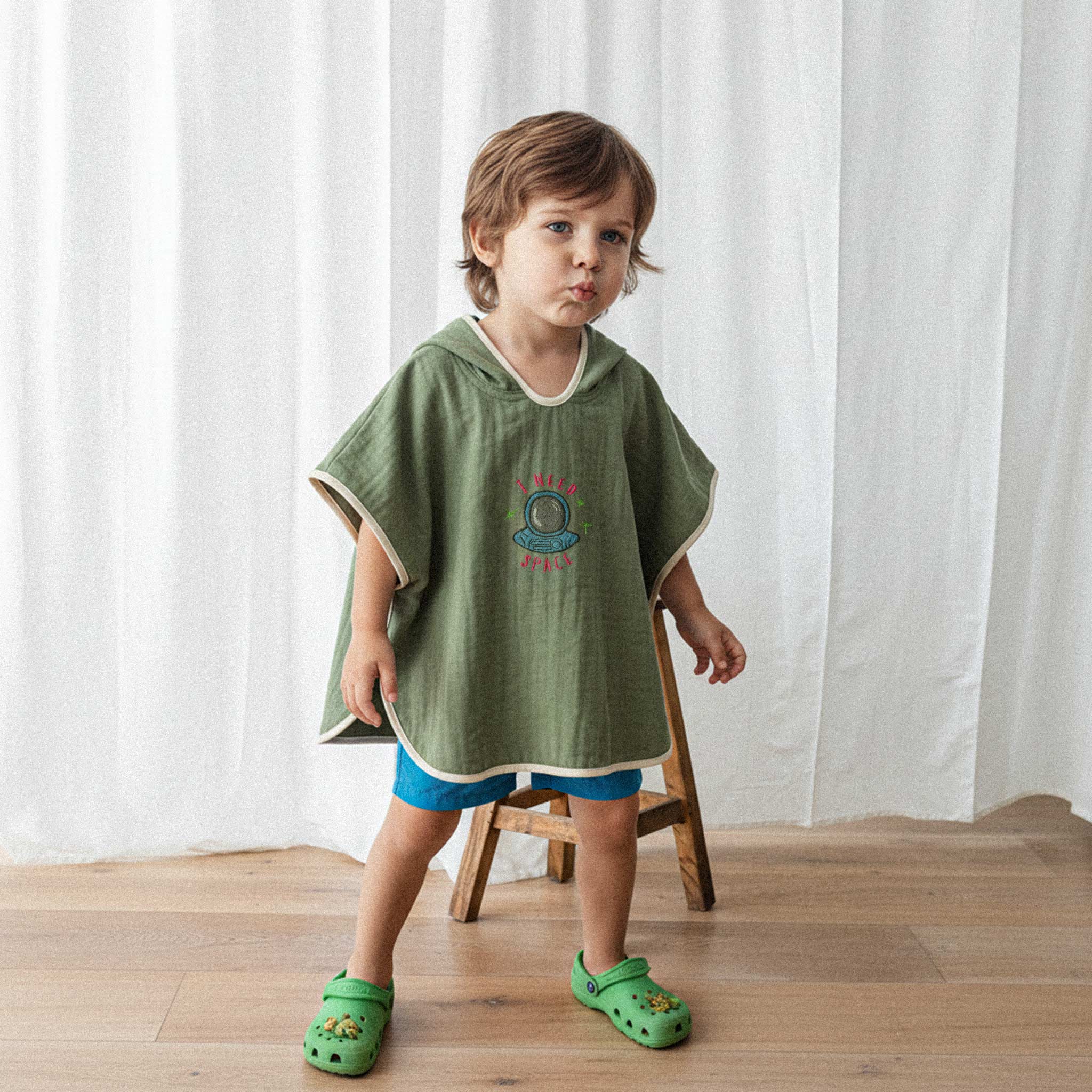 Mavi şort ve haki yeşili, 'I Need Space' astronot nakışlı kapüşonlu müslin panço giyen erkek çocuk, havalı bir poz vererek kameraya bakıyor.
