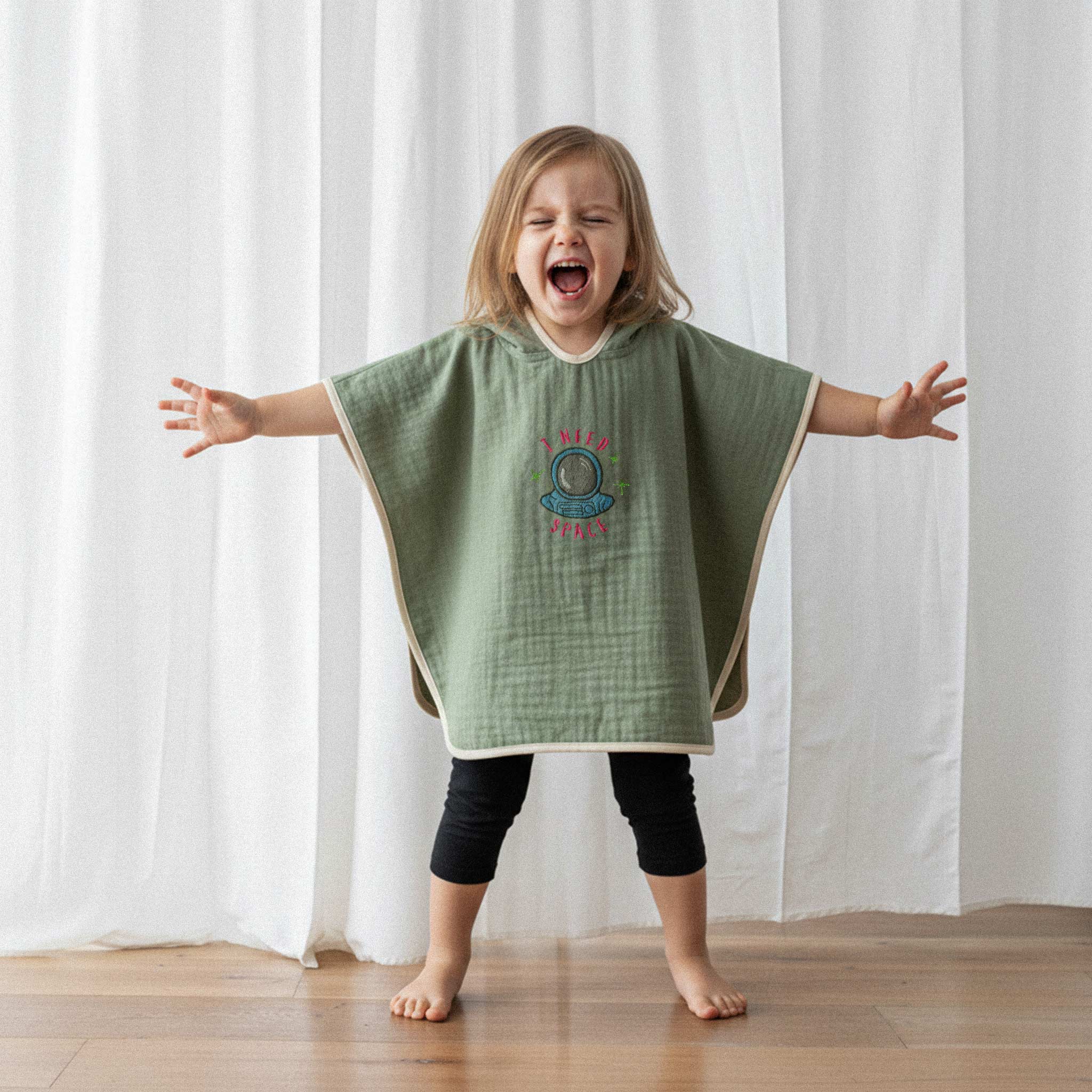Siyah tayt ve haki yeşili, 'I Need Space' astronot nakışlı kapüşonlu müslin panço giyen küçük kız çocuk, kollarını açarak neşeyle bağırıyor.