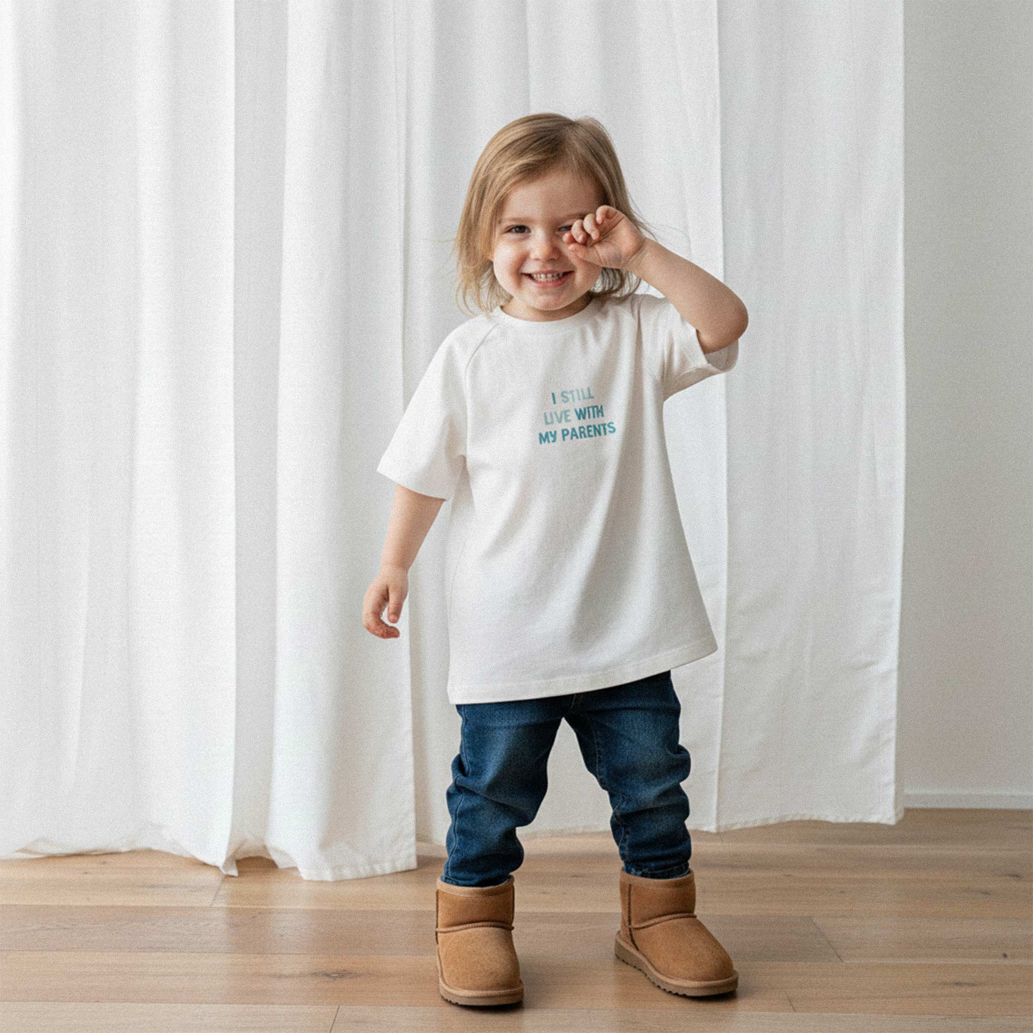 Jean pantolon ve ekru renkli, 'I Still Live With My Parents' nakışlı reglan kollu oversize bebek tişörtü giyen küçük kız çocuk, gözünü ovuşturarak tatlı bir şekilde gülümsüyor.