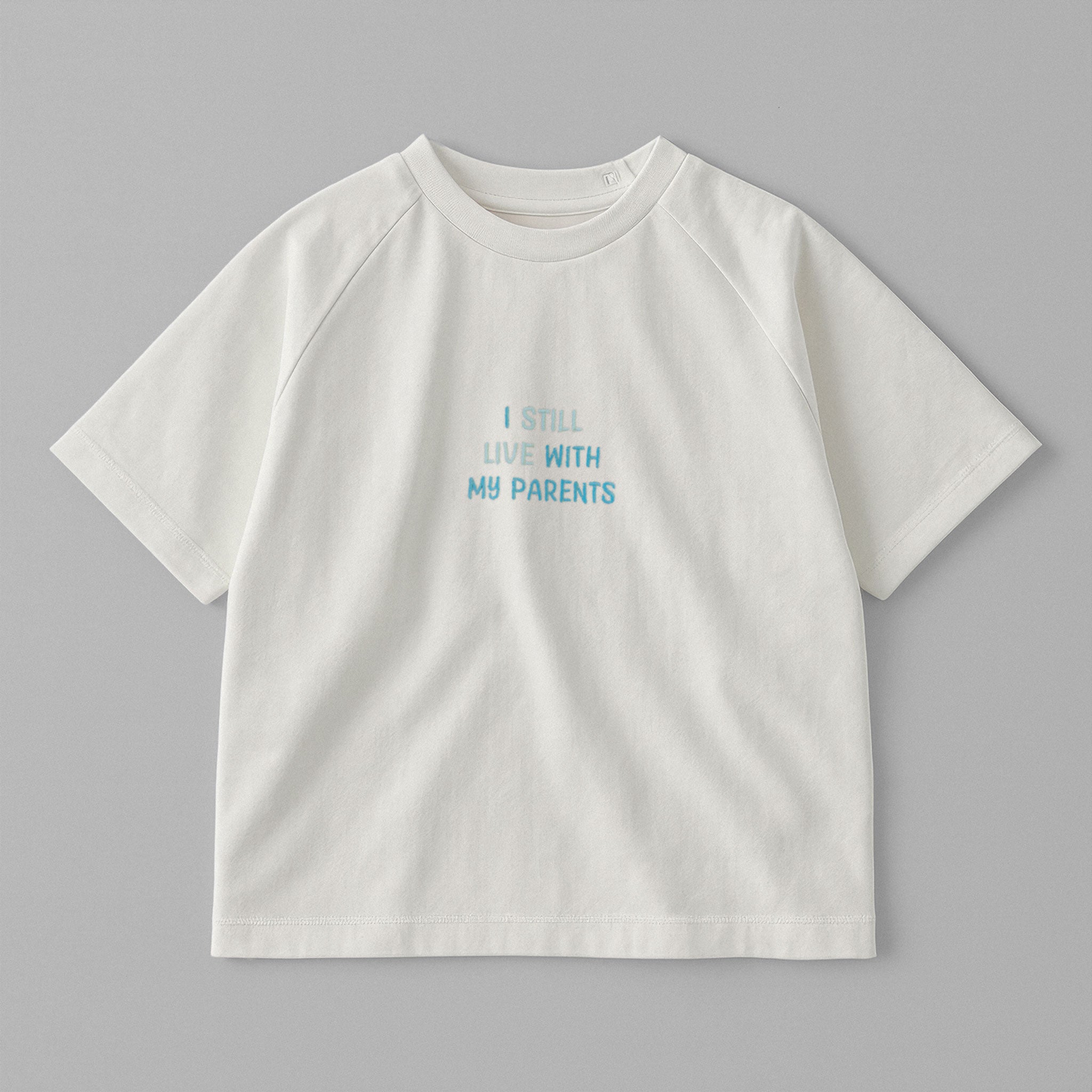 Ekru renkli, reglan kollu, omuzdan çıtçıtlı bebek tişörtü. Göğüs kısmında mavi tonlarda "I STILL LIVE WITH MY PARENTS" nakışı bulunmaktadır.