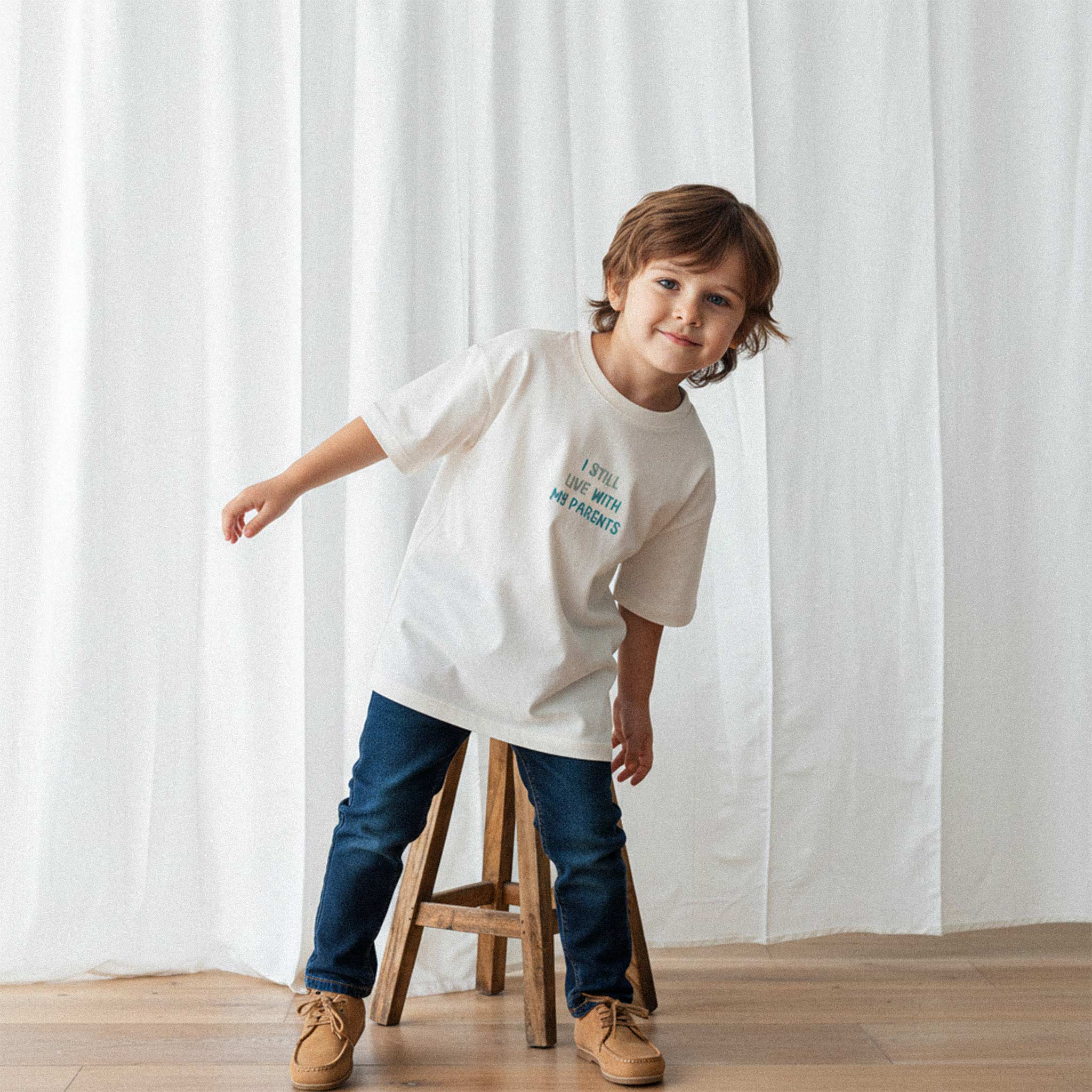 Jean pantolon ve ekru renkli, 'I Still Live With My Parents' nakışlı oversize tişört giyen erkek çocuk, ahşap bir tabureye dayanarak sevimli bir poz veriyor.