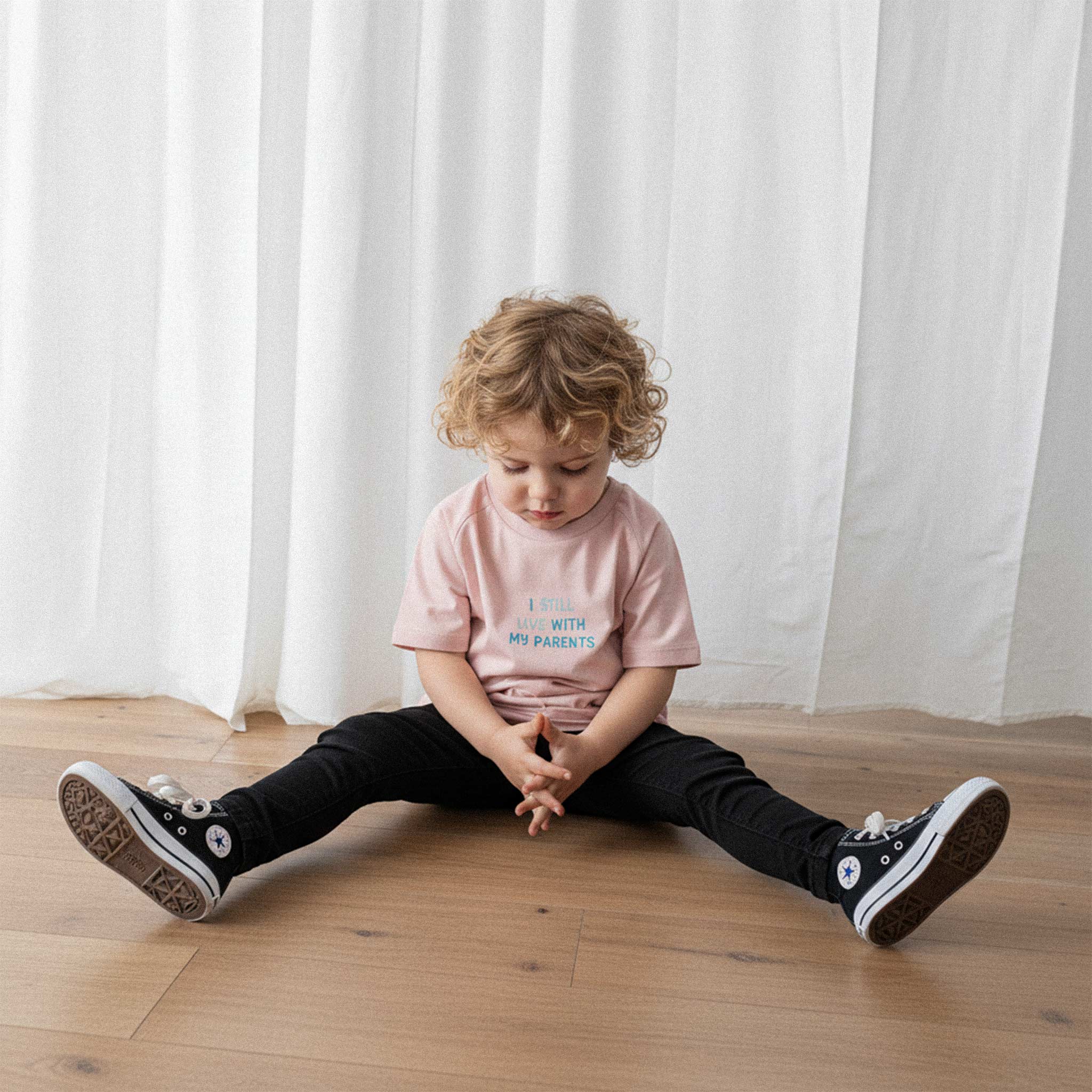 Siyah pantolon ve gül kurusu, 'I Still Live With My Parents' nakışlı oversize tişört giyen kıvırcık saçlı küçük erkek çocuk, yerde otururken düşünceli bir şekilde tişörtüne bakıyor.