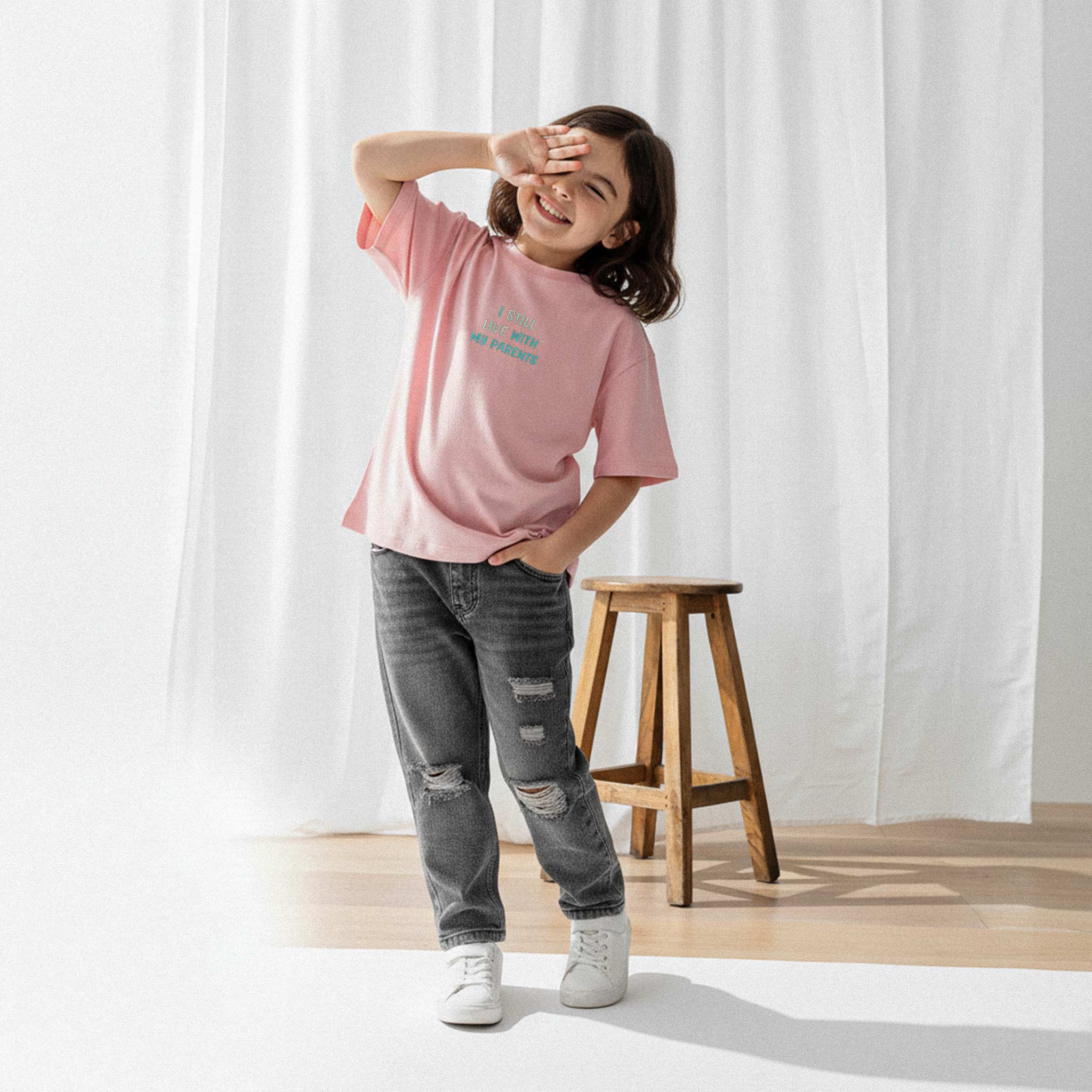 Yırtık gri jean ve gül kurusu, 'I Still Live With My Parents' nakışlı oversize tişört giyen kız çocuk, eliyle gözünü güneşten koruyarak gülümsüyor.