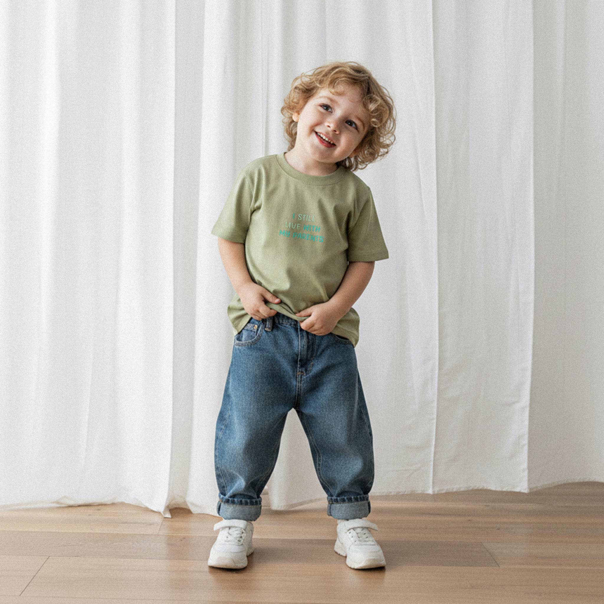 Bol paça jean pantolon ve haki yeşili, 'I Still Live With My Parents' nakışlı oversize tişört giyen kıvırcık saçlı küçük erkek çocuk, sevimli bir şekilde gülümseyerek poz veriyor.