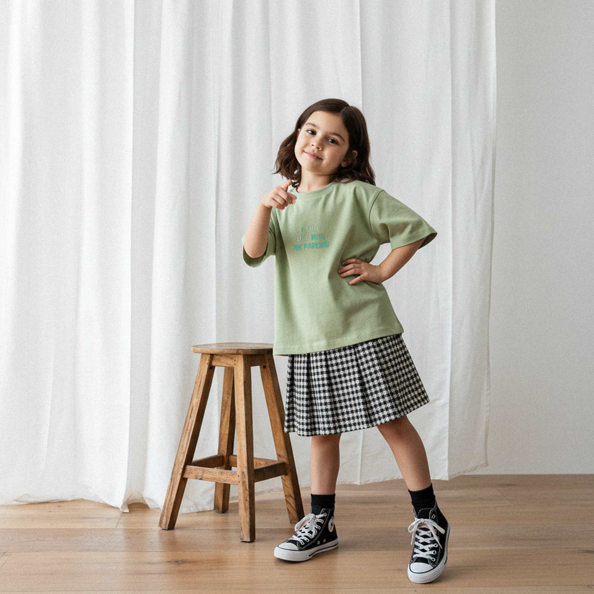 Pötikare etek ve haki yeşili, 'I Still Live With My Parents' nakışlı oversize tişört giyen kız çocuk, elini beline koyarak havalı bir poz veriyor.