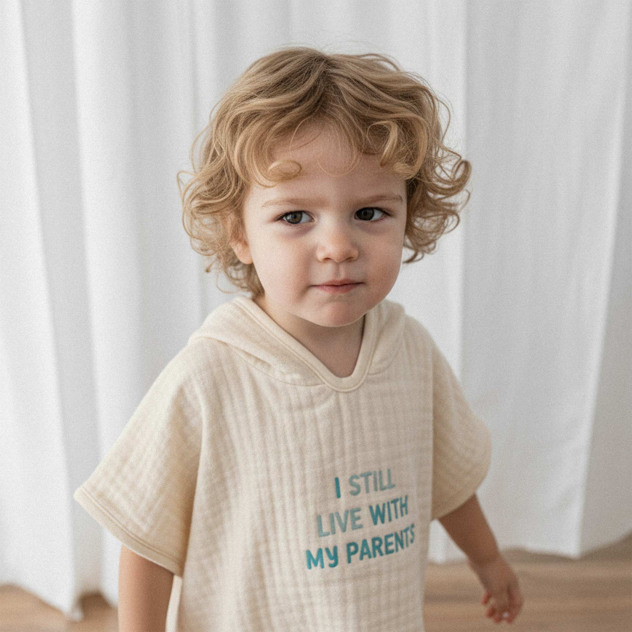 Üzerinde ekru renkli, 'I Still Live With My Parents' nakışlı kapüşonlu müslin panço bulunan kıvırcık saçlı küçük erkek çocuğun sevimli yakın çekim portresi.