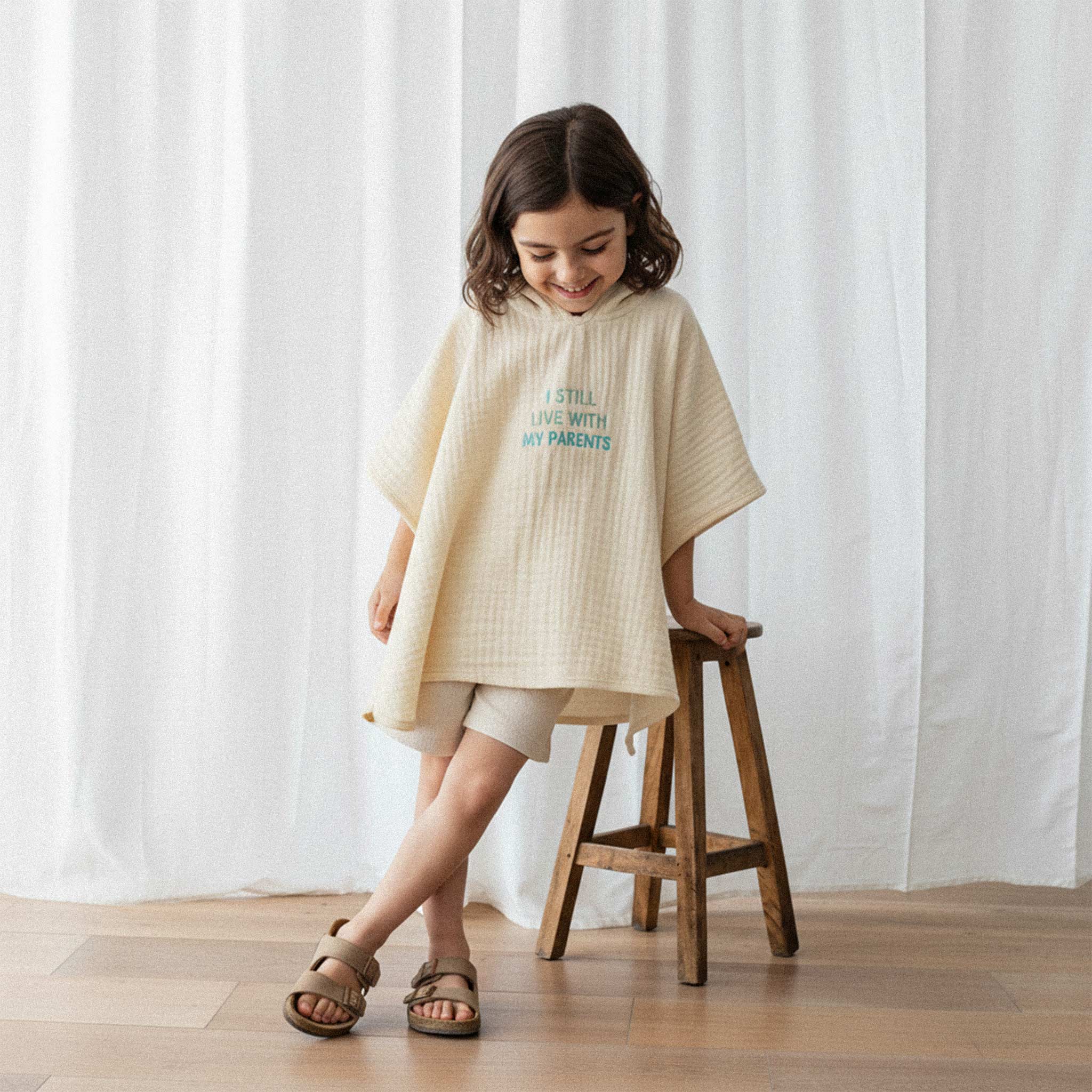 Bej şort ve sandalet giyen kız çocuk, üzerinde ekru renkli, 'I Still Live With My Parents' nakışlı kapüşonlu müslin panço ile ahşap bir tabureye yaslanarak gülümsüyor.