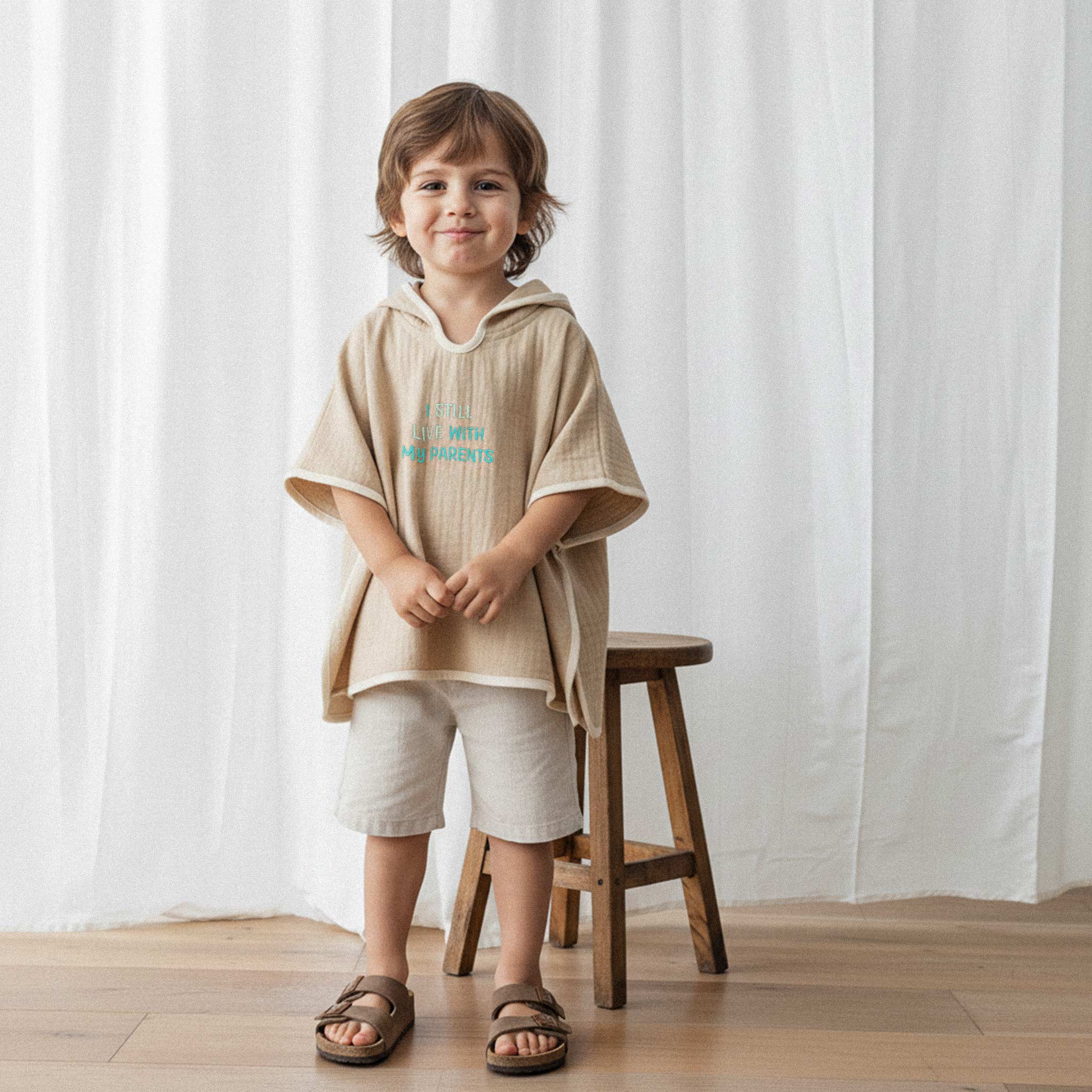 Keten şort ve somon rengi, 'I Still Live With My Parents' nakışlı kapüşonlu müslin panço giyen erkek çocuk, gülümseyerek poz veriyor.