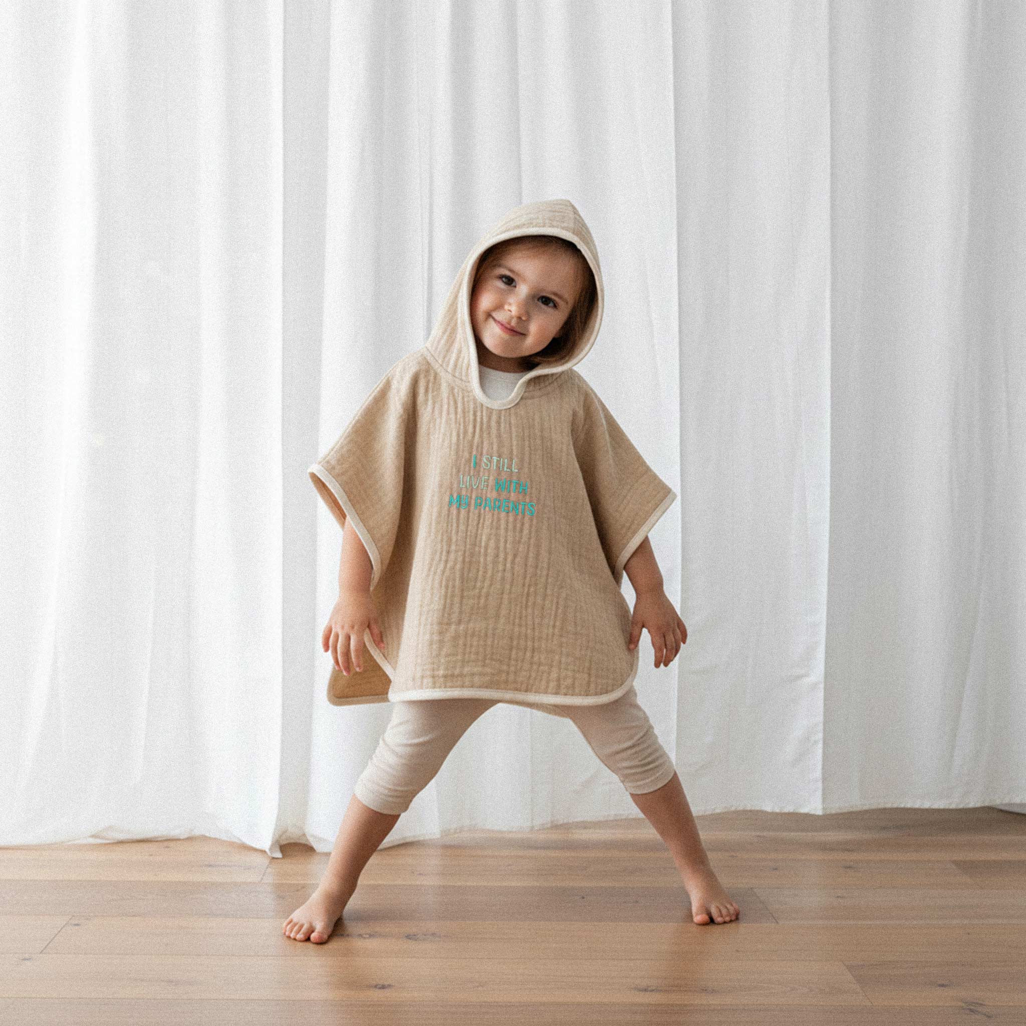 Bej tayt ve somon rengi, 'I Still Live With My Parents' nakışlı kapüşonlu müslin panço giyen küçük kız çocuk, gülümseyerek eğlenceli bir poz veriyor.