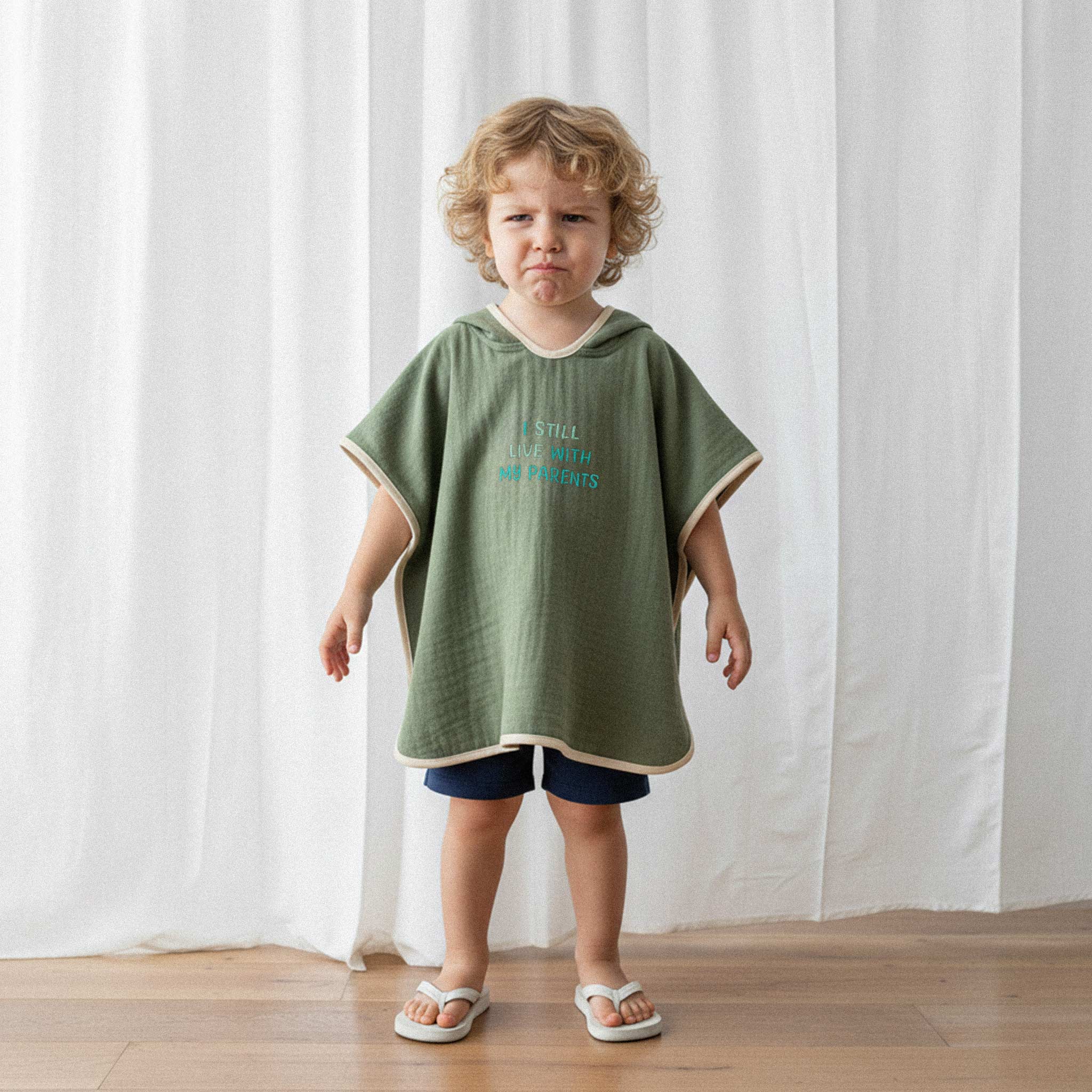 Lacivert şort ve haki yeşili, 'I Still Live With My Parents' nakışlı kapüşonlu müslin panço giyen kıvırcık saçlı küçük erkek çocuk, komik bir surat ifadesiyle poz veriyor.