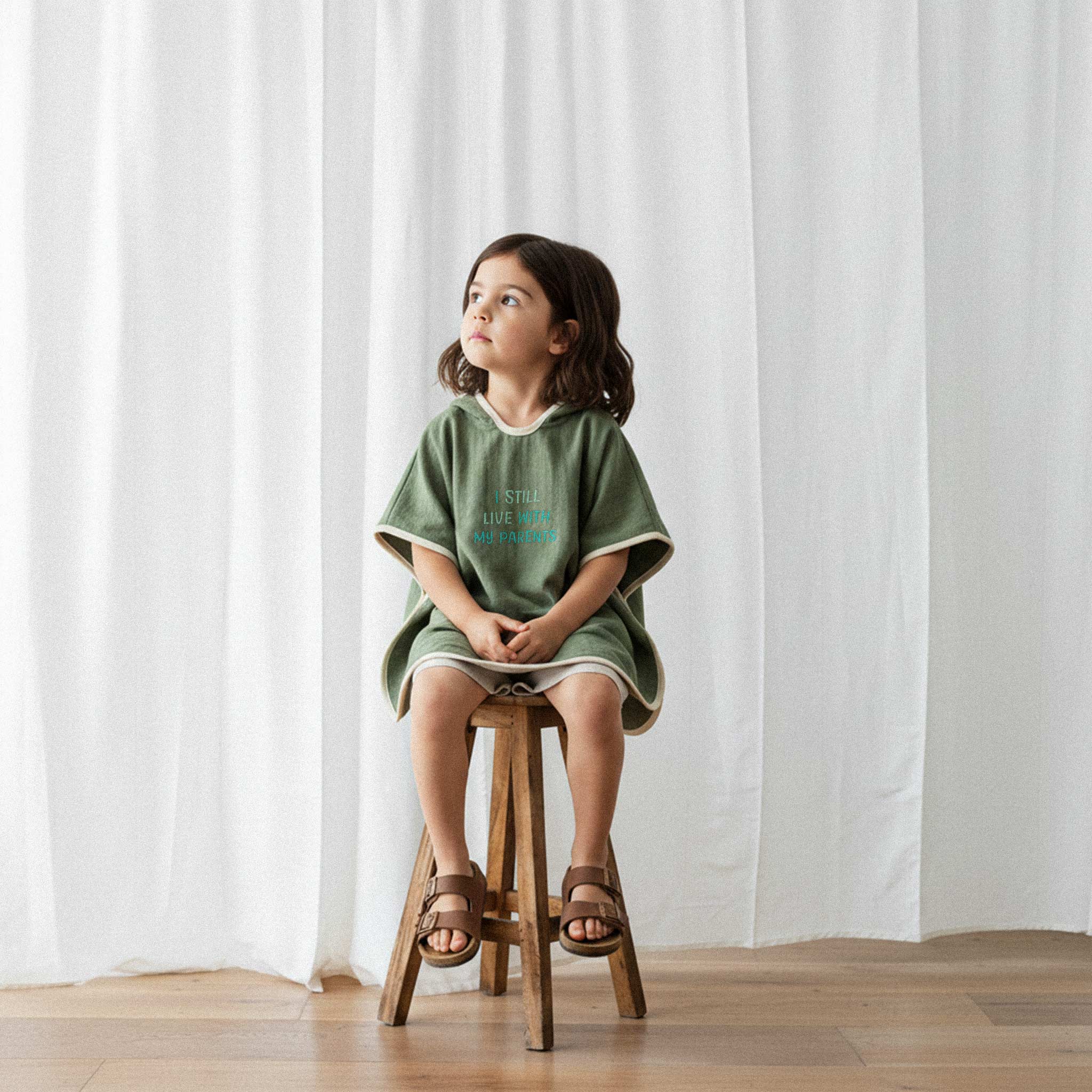 Elbise üzerine haki yeşili, 'I Still Live With My Parents' nakışlı kapüşonlu müslin panço giyen kız çocuk, ahşap bir taburede otururken düşünceli bir şekilde yana bakıyor.