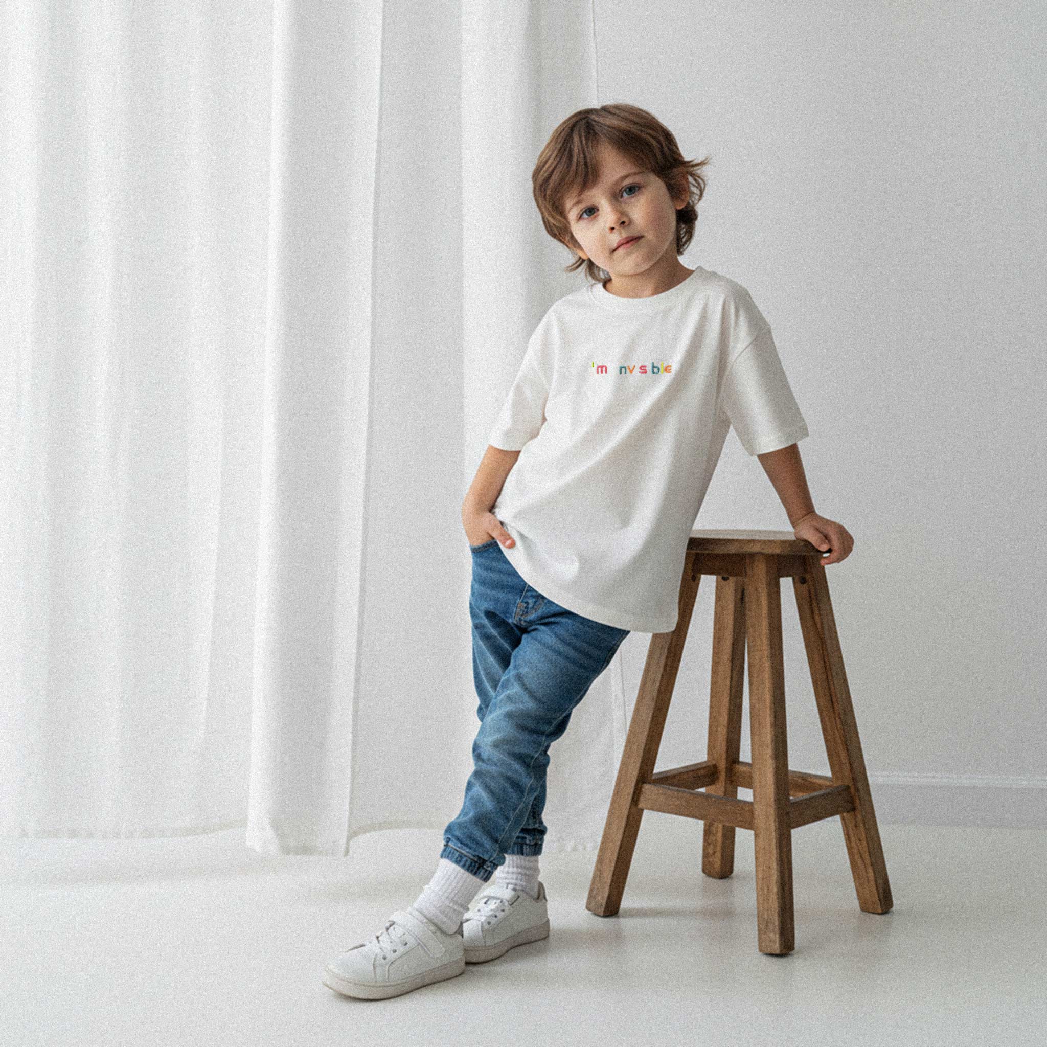 Jean pantolon ve ekru renkli, 'I'm nvsble' nakışlı oversize tişört giyen erkek çocuk, ahşap bir tabureye yaslanarak havalı bir poz veriyor.