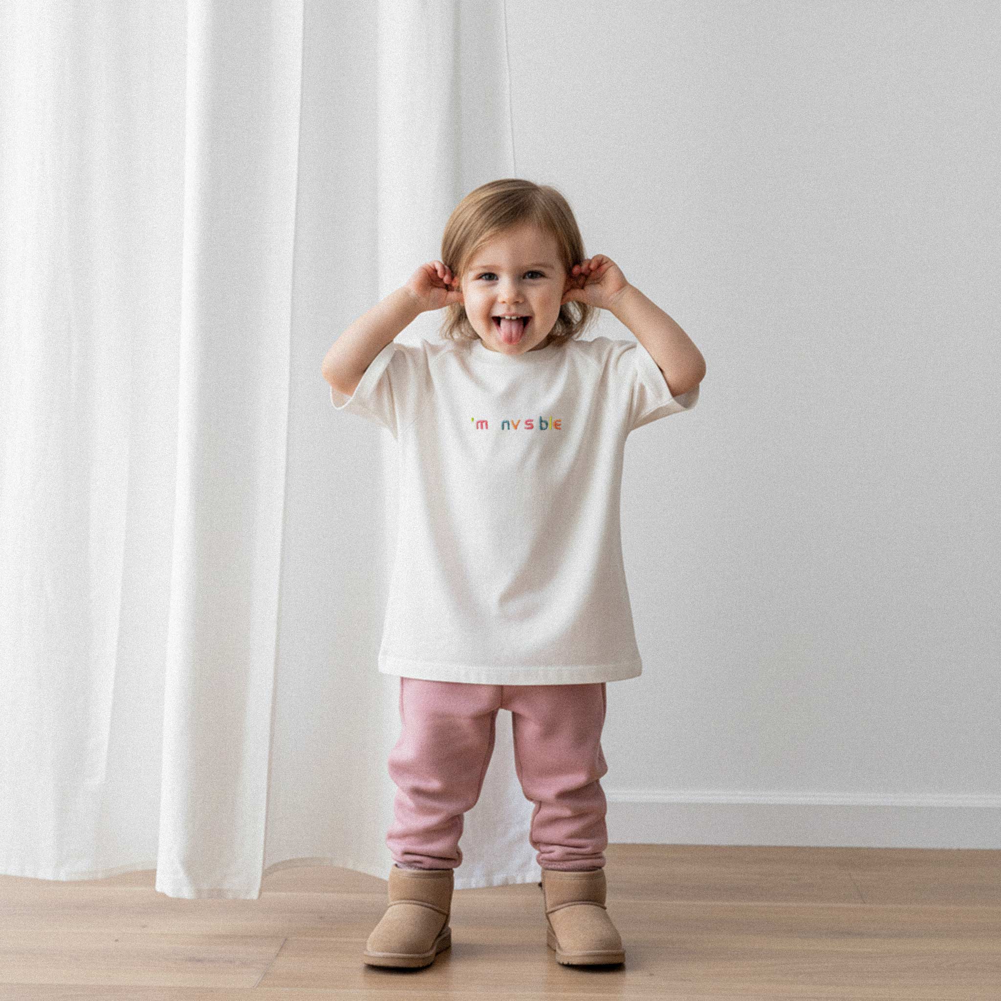 Pembe eşofman altı ve ekru renkli, 'I'm nvsble' nakışlı oversize tişört giyen küçük kız çocuk, dilini çıkarıp kulaklarını çekerek komik bir surat ifadesi yapıyor.
