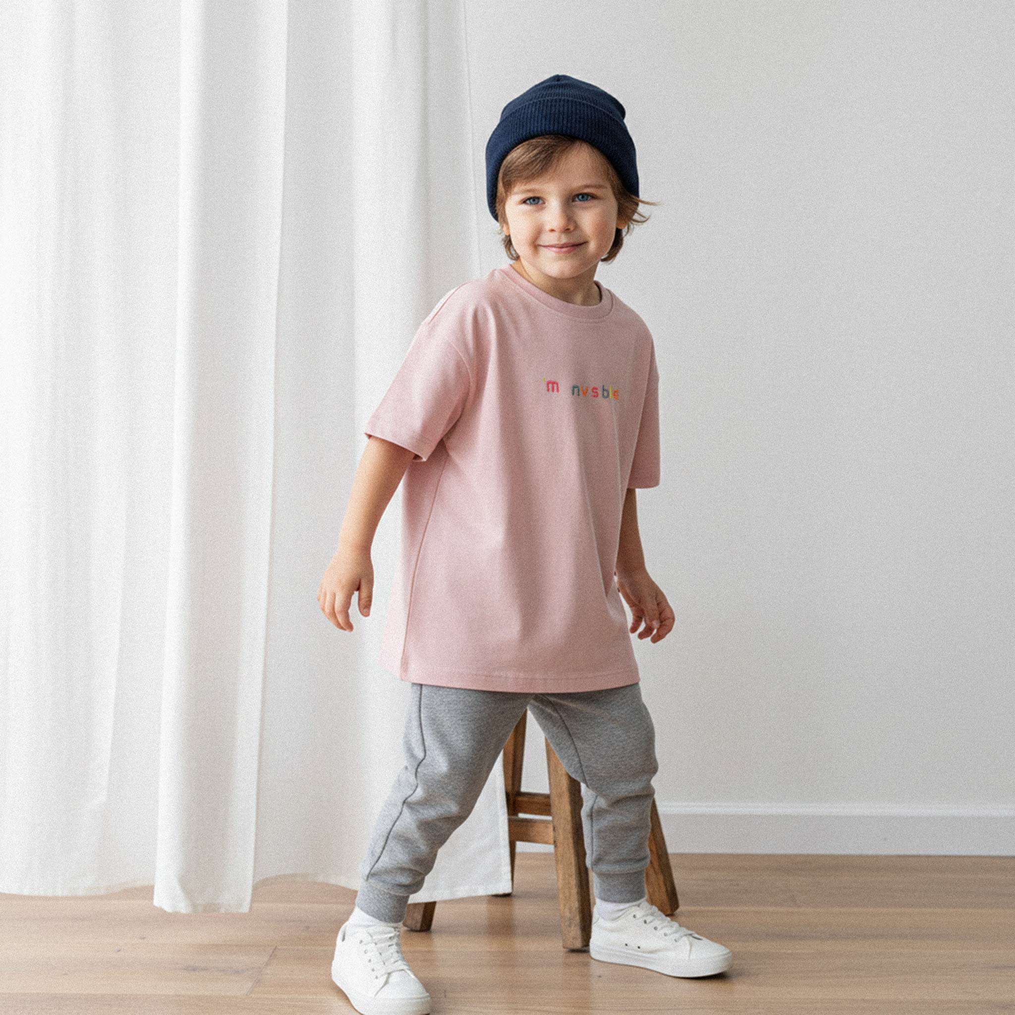 Mavi bere takan, gri eşofman altı ve gül kurusu, 'I'm nvsble' nakışlı oversize tişört giyen erkek çocuk, gülümseyerek poz veriyor.