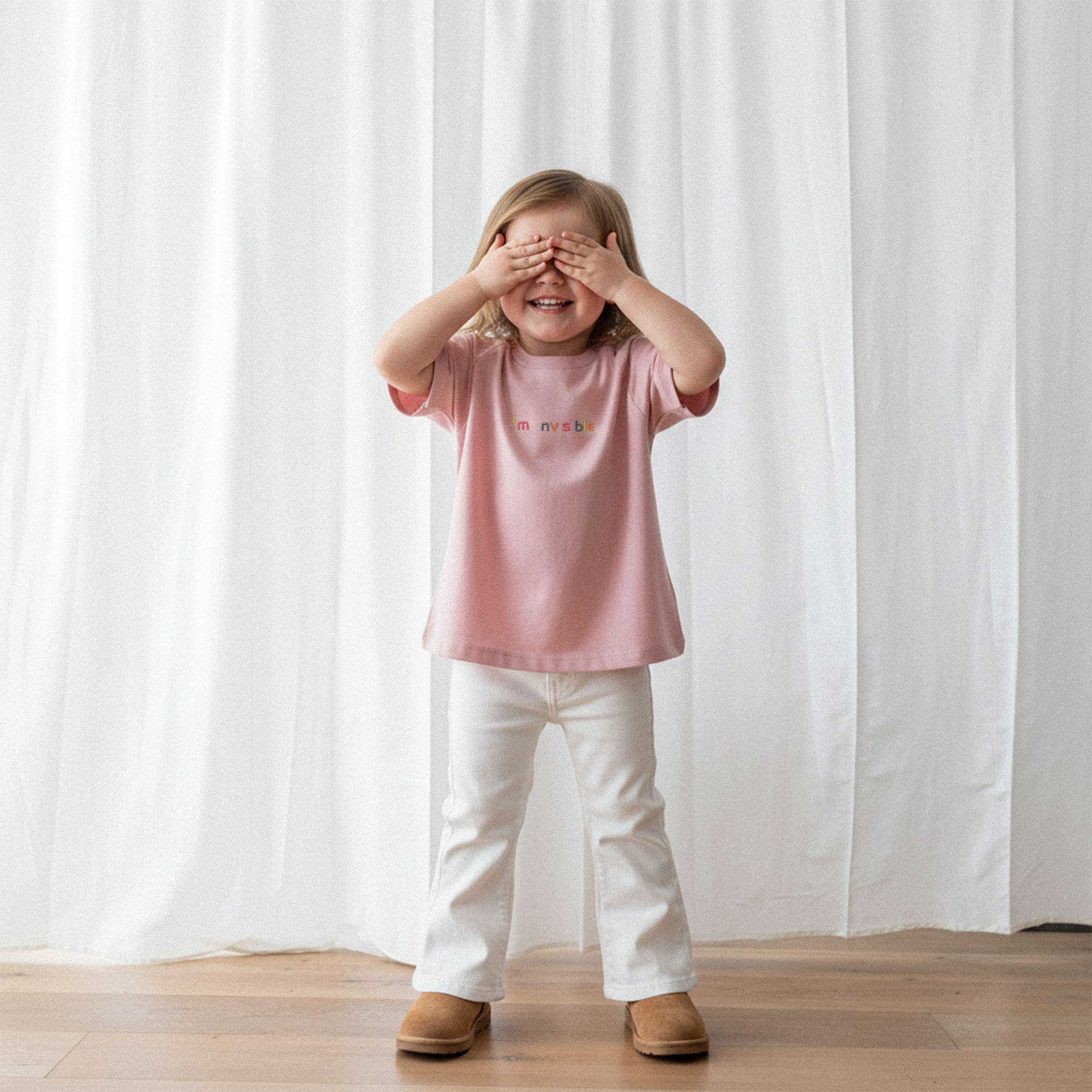 Beyaz pantolon ve gül kurusu, 'I'm nvsble' nakışlı oversize tişört giyen küçük kız çocuk, gülümseyerek elleriyle gözlerini kapatıyor.