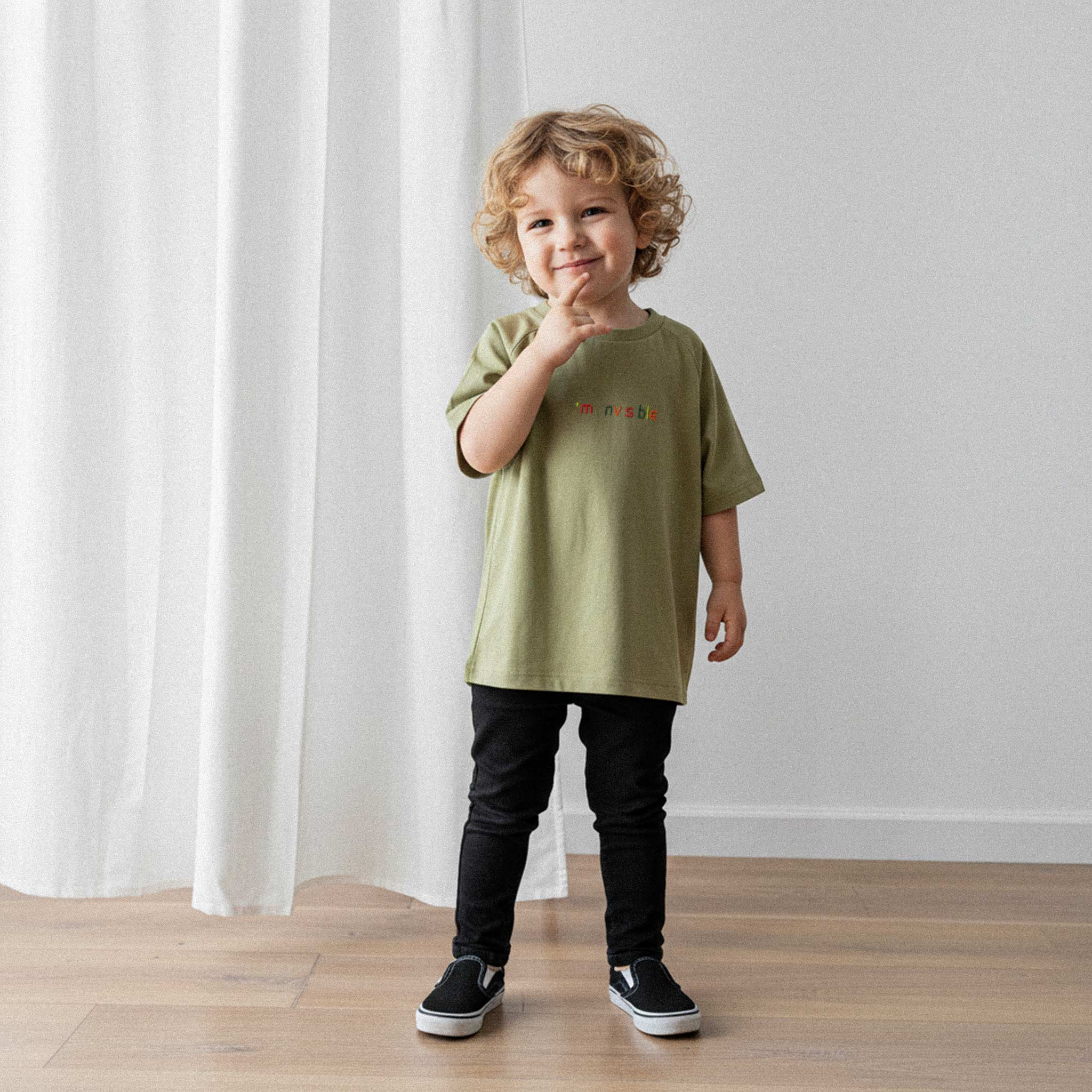 Siyah pantolon ve haki yeşili, 'I'm nvsble' nakışlı oversize tişört giyen kıvırcık saçlı erkek çocuk, parmağı çenesinde sevimli bir poz veriyor.