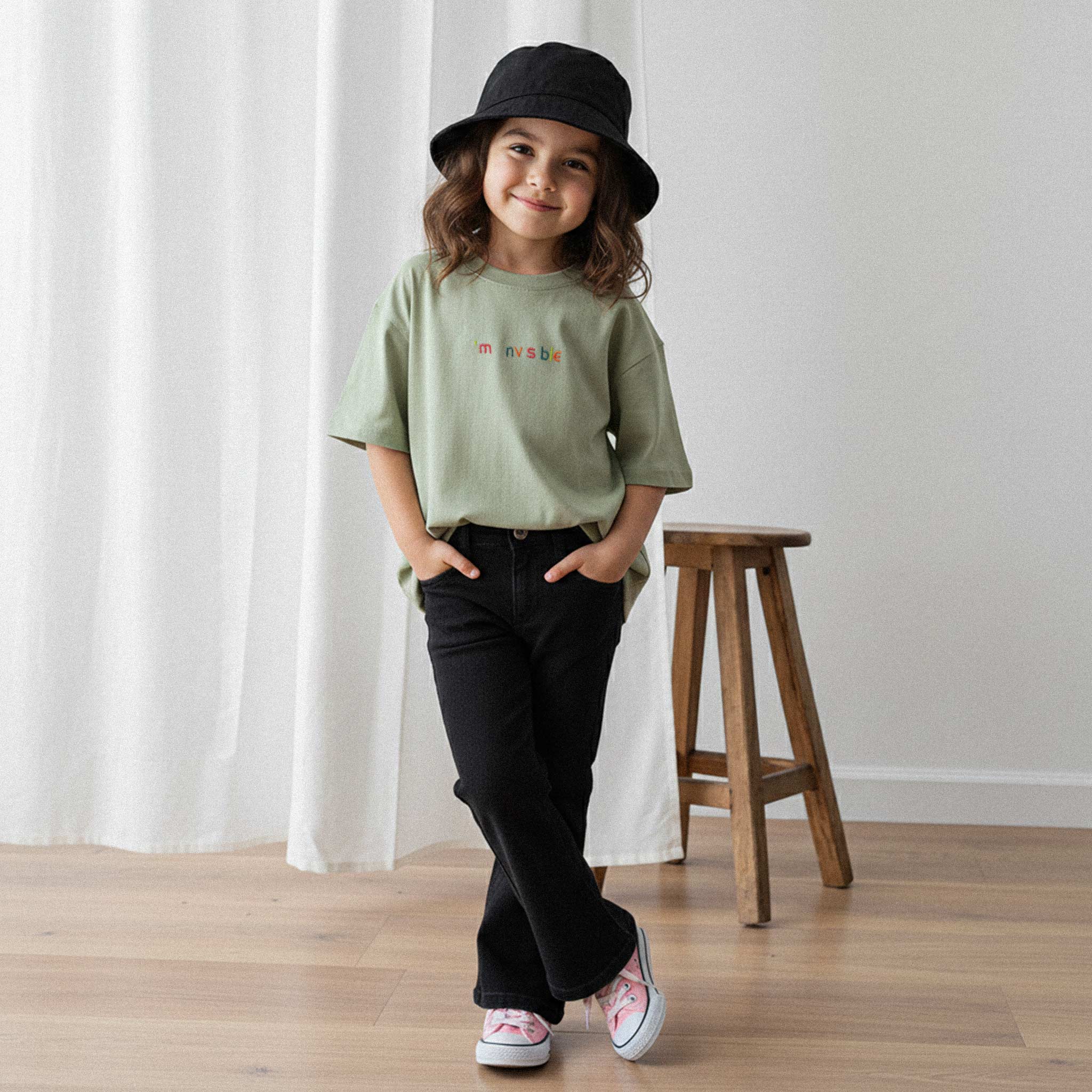 Siyah pantolon, bucket şapka ve haki yeşili, 'I'm nvsble' nakışlı oversize tişört giyen kız çocuk, elleri ceplerinde gülümseyerek poz veriyor.
