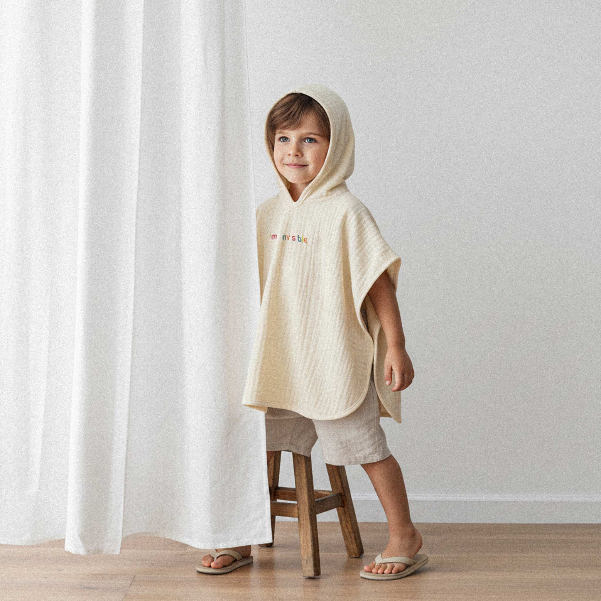 Bej şort ve terlik giyen erkek çocuk, üzerinde ekru renkli, 'I'm nvsble' nakışlı kapüşonlu müslin panço ile gülümseyerek poz veriyor.