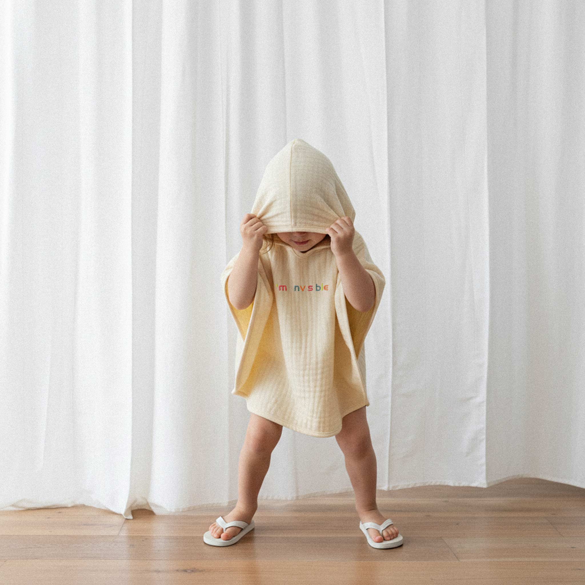 Üzerinde ekru renkli, 'I'm nvsble' nakışlı kapüşonlu müslin panço bulunan küçük çocuk, kapüşonunu yüzüne çekerek "görünmez" olma oyunu oynuyor.