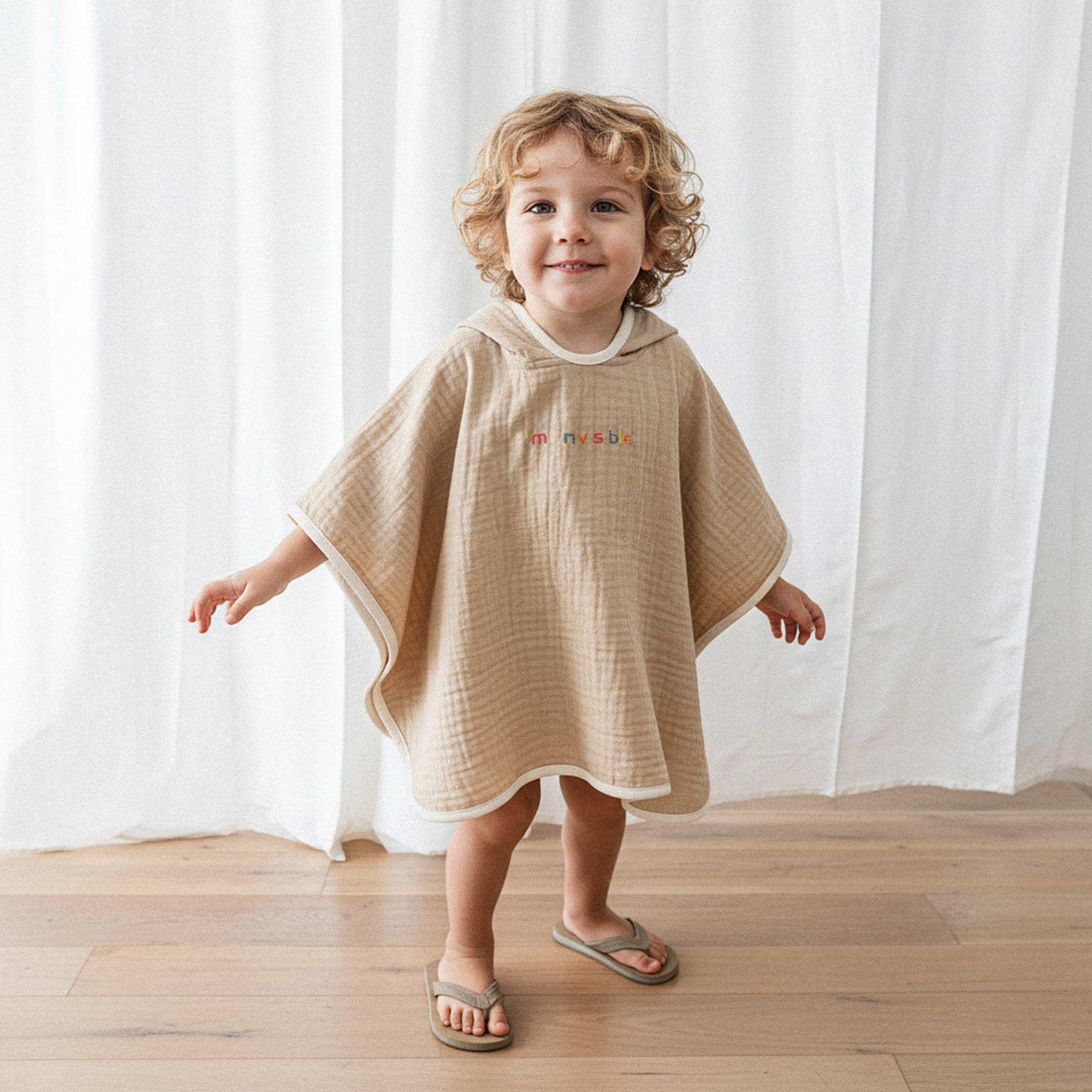 Kıvırcık saçlı küçük erkek çocuk, üzerinde somon rengi, 'I'm nvsble' nakışlı kapüşonlu müslin panço ile kollarını açarak neşeyle gülümsüyor.
