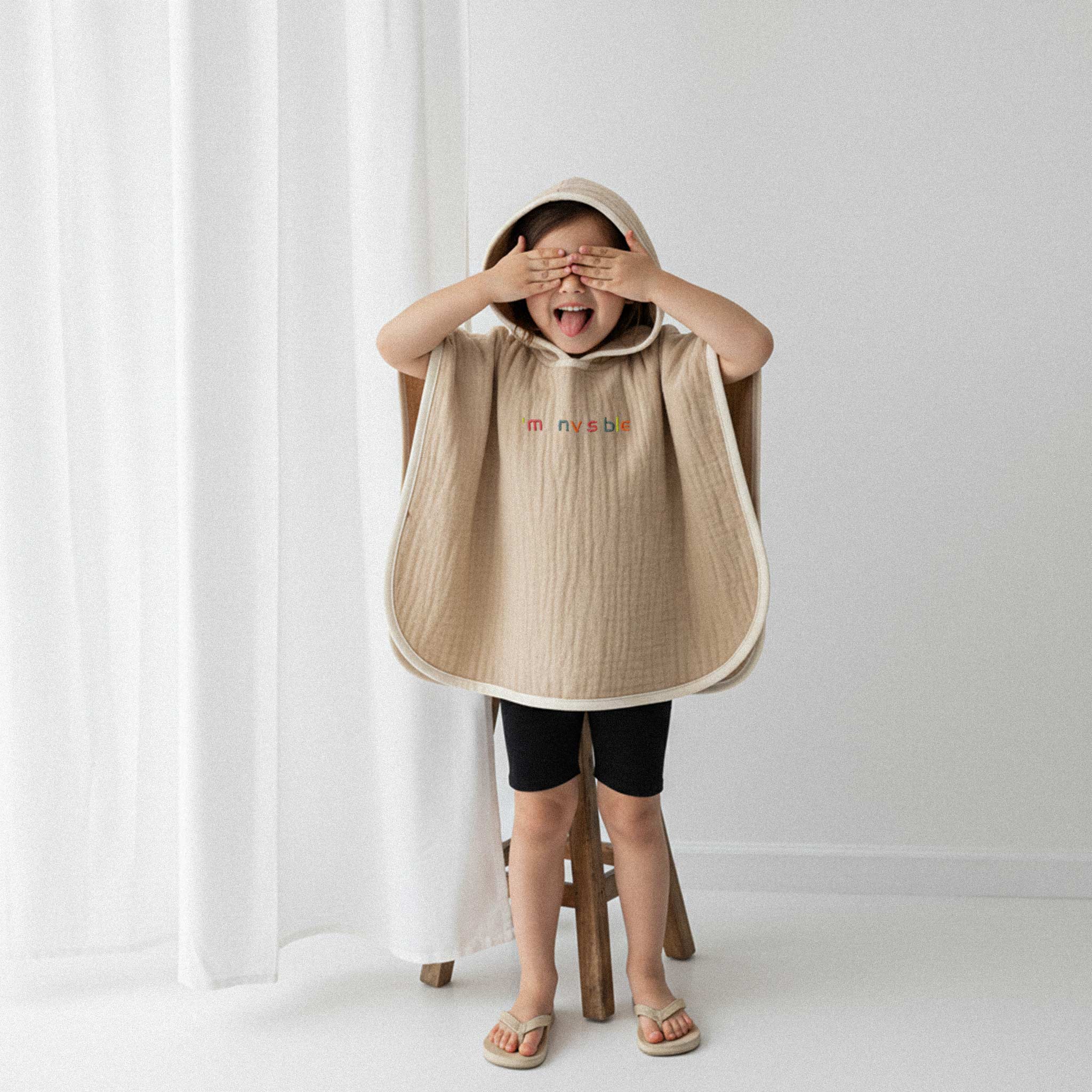 Üzerinde somon rengi, 'I'm nvsble' nakışlı kapüşonlu müslin panço bulunan küçük kız çocuk, sandalyede dururken elleriyle gözlerini kapatarak eğlenceli bir poz veriyor.