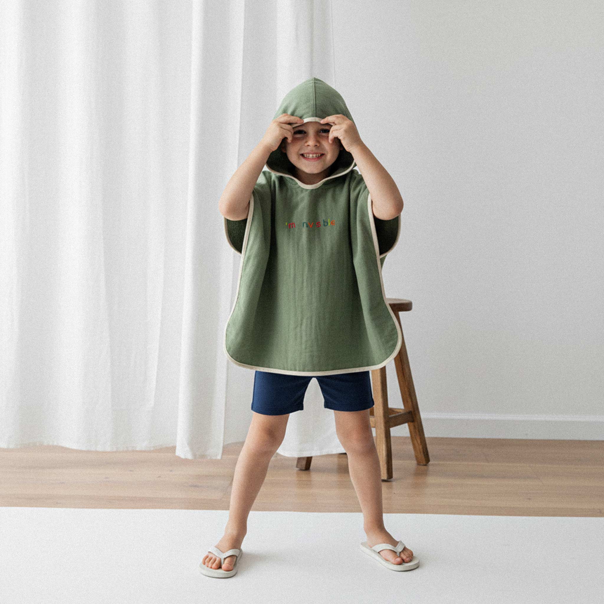 Lacivert şort ve haki yeşili, 'I'm nvsble' nakışlı kapüşonlu müslin panço giyen erkek çocuk, elleriyle gözlerini tutarak eğlenceli bir poz veriyor.