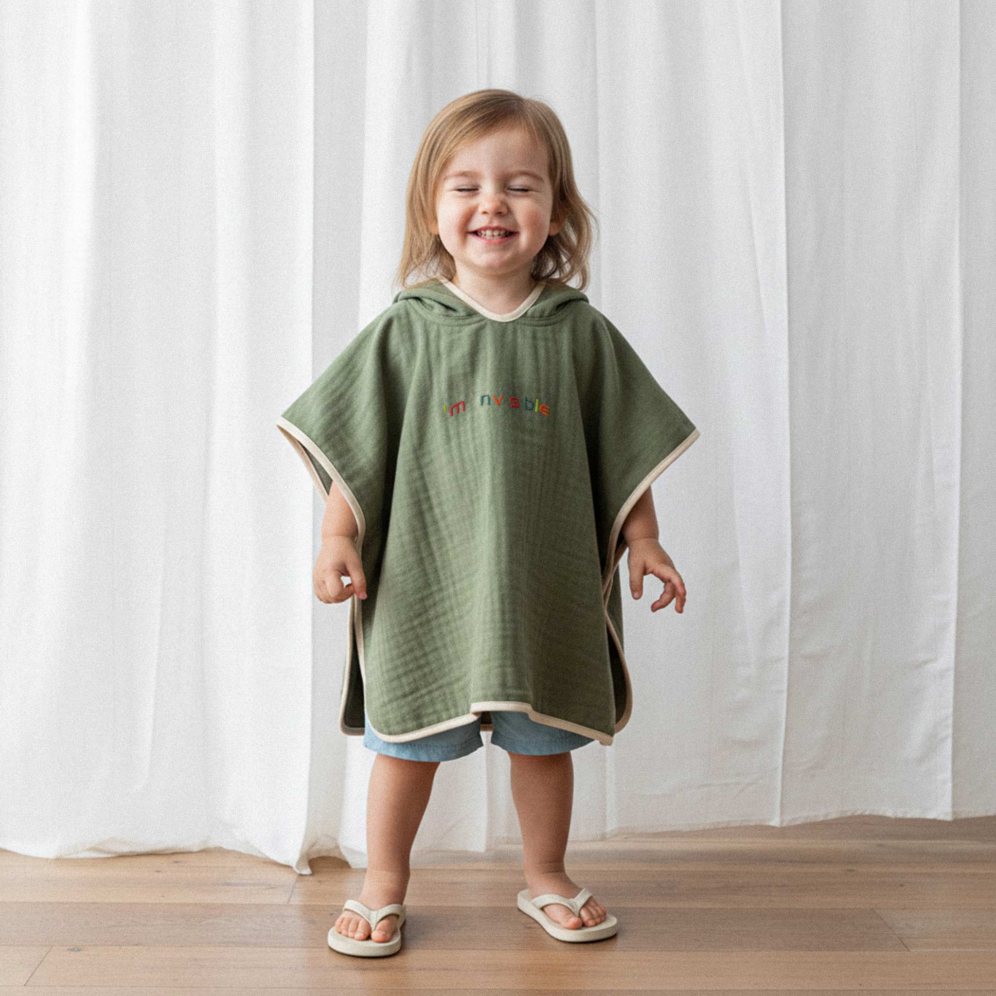 Üzerinde haki yeşili renkli, 'I'm nvsble' nakışlı kapüşonlu müslin panço bulunan küçük kız çocuk, neşeyle gözleri kapalı gülümseyerek poz veriyor.