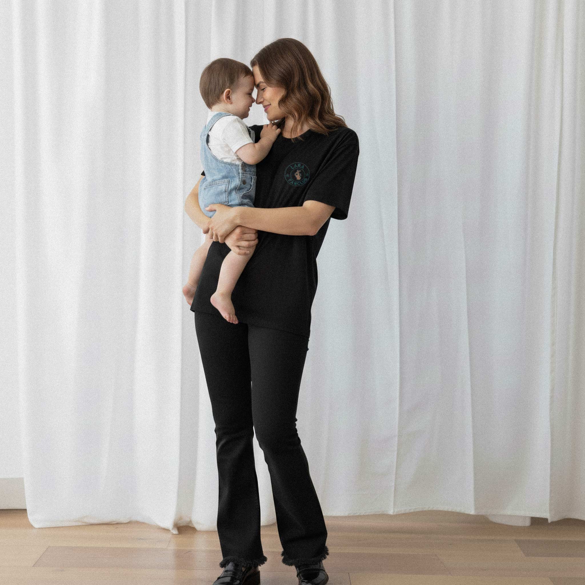 Kucağındaki bebeğe sarılan anne, üzerinde siyah renkli, kişiye özel 'LARA Fanclub' nakışlı oversize Bona Pera tişörtü giyiyor.