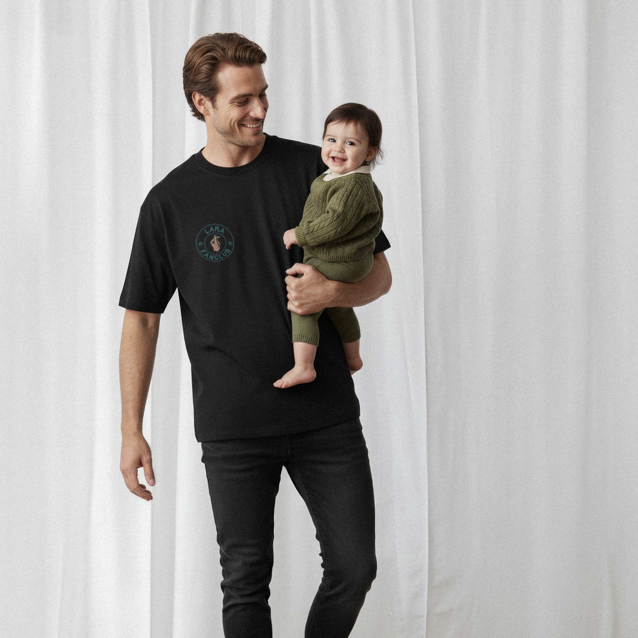 Kucağındaki bebeğiyle gülümseyen baba, üzerinde siyah renkli, kişiye özel 'LARA Fanclub' nakışlı oversize Bona Pera tişörtü giyiyor.