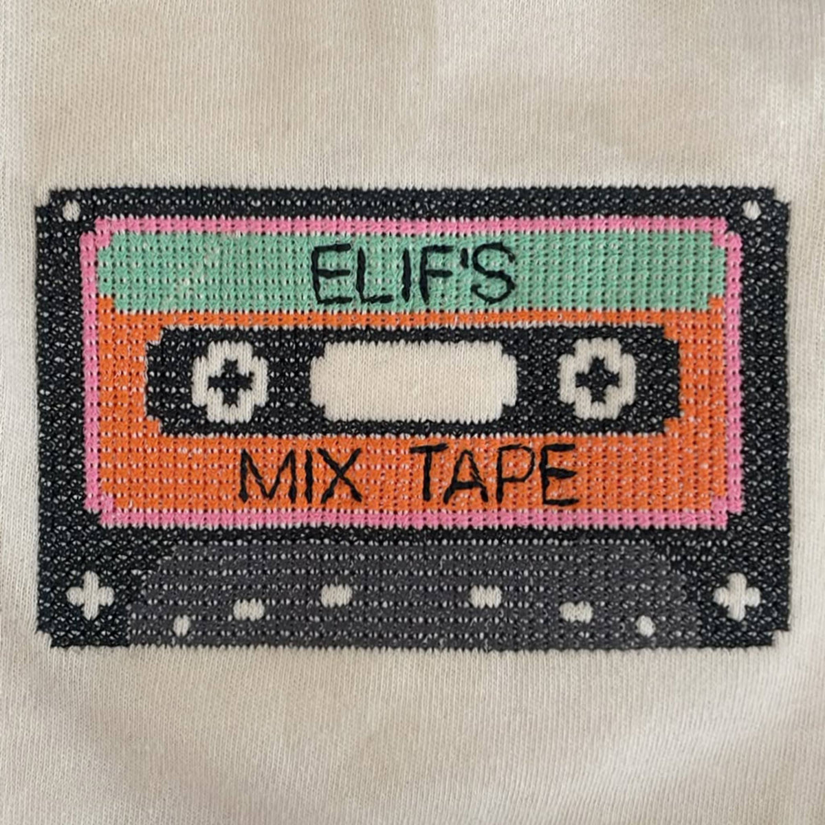 Ekru sweatshirt üzerine cross stitch (çarpı işi) tekniğiyle işlenmiş, 70'ler ve 80'lerin nostaljik sembolü kaset (mix tape) nakışının yakından görünümü. Pixel art temalı bu renkli tasarım, görseldeki gibi üzerine dilediğiniz isim yazılarak tamamen kişiselleştirilebilir. Birinci sınıf Alman nakış ipliği kullanılarak yüksek kalite standartlarında, ince bir işçilikle üretilmiştir. Çocuklar için eğlenceli ve isme özel premium nakış detayı.
