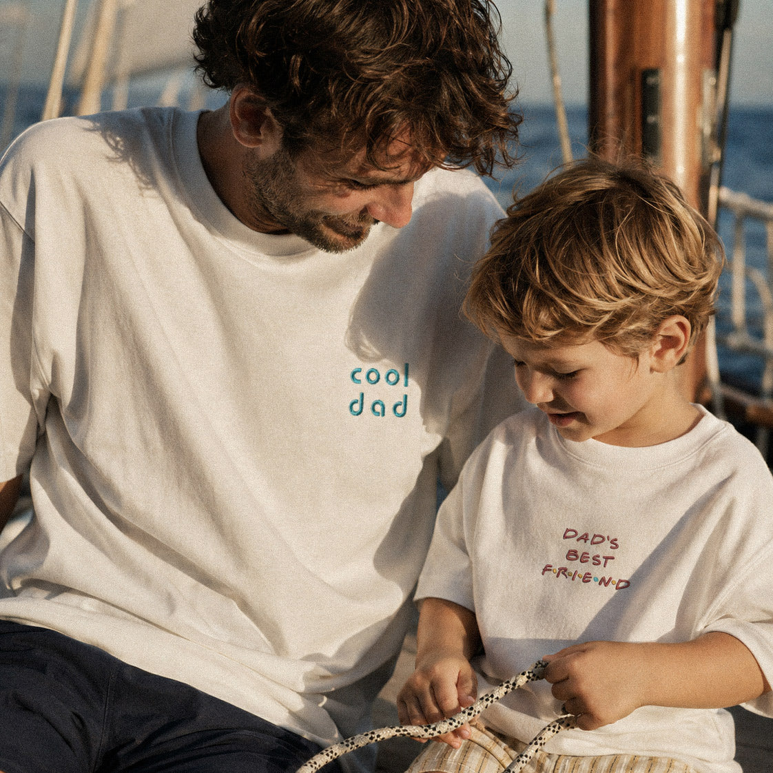 Teknede baba-oğul lifestyle çekimi. Babada mavi "cool dad" nakışlı, çocukta "DAD'S BEST F.R.I.E.N.D" nakışlı kırık beyaz %100 pamuklu oversize tişört. Birinci sınıf Alman işçiliği yazlık premium aile kombini.