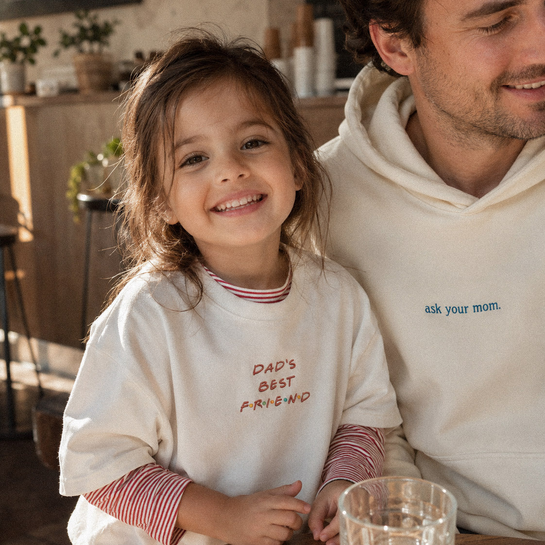 Kırık beyaz renkli, ikonik dizi fontuyla renkli iplikle işlenmiş "DAD'S BEST FRIEND" nakışlı %100 pamuklu çocuk tişörtü ile mavi iplikle "ask your mom." işlenmiş krem rengi Oeko-Tex sertifikalı içi şardonlu baba kapüşonlu sweatshirt kombini. Birinci sınıf Alman nakış işçiliğiyle üretilmiş ebeveyn-çocuk uyumunu yansıtan premium lifestyle ürün çekimi.
