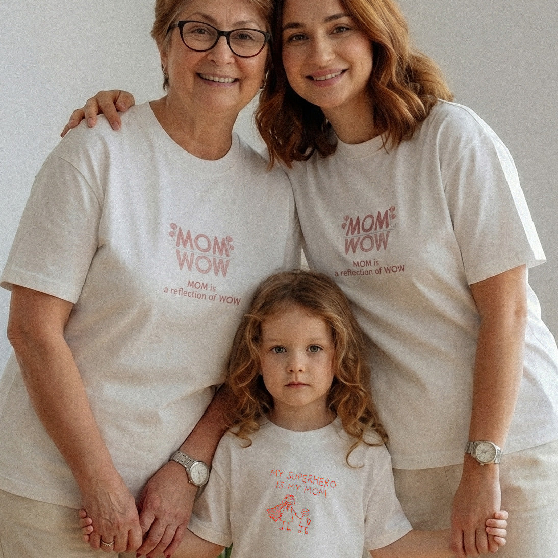 Üç kuşak lifestyle çekimi. Anneanne ve annede kırık beyaz %100 pamuklu, pembe "MOM WOW" nakışlı oversize tişört; çocukta "MY SUPERHERO IS MY MOM" ve pelerinli anne nakışlı uyumlu tişört. Birinci sınıf Alman işçiliği premium aile kombini.