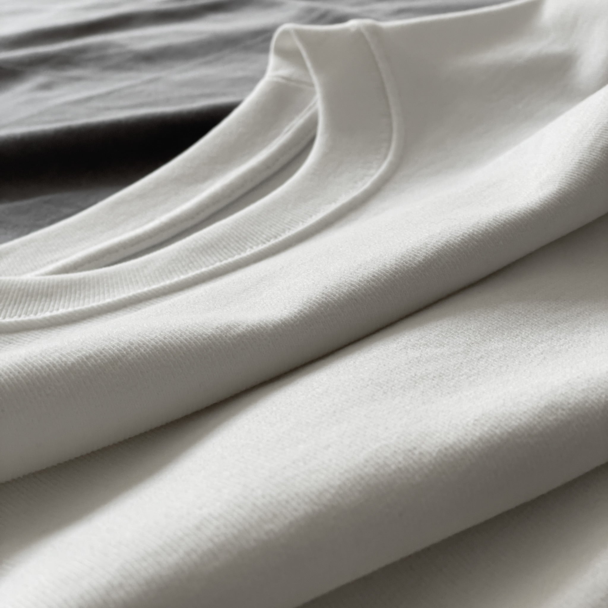 Kırık beyaz (ekru) premium yetişkin t-shirt modelinin tok pamuklu kumaş dokusunu ve yaka kısmını gösteren katlanmış ürün çekimi. Ağır gramajlı (heavyweight) kumaşın yoğun örgüsü dökümlü kesimin formunu korur ve tok bir duruş sağlar. Dayanıklılık ve premium hissi vurgulayan görsel.