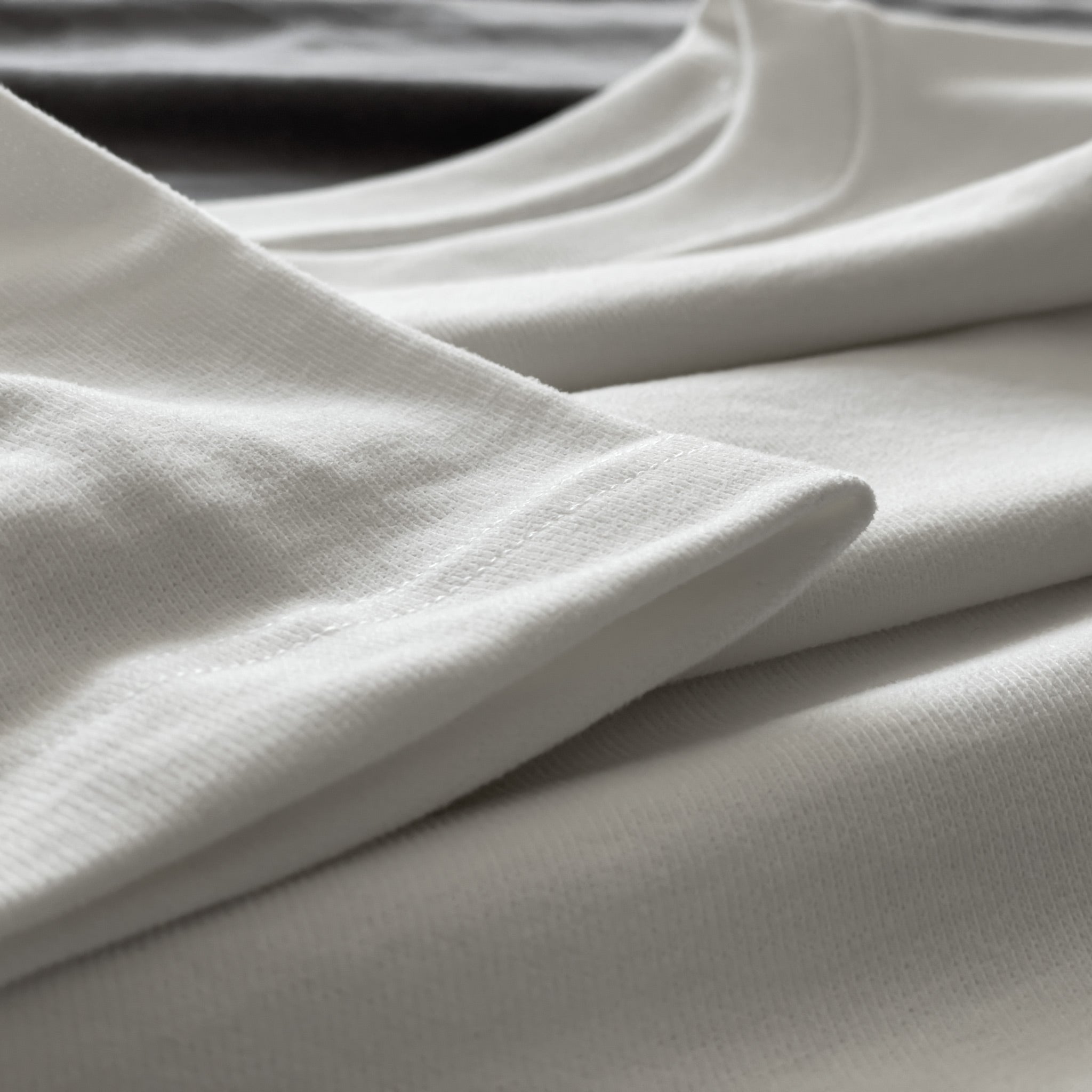 Kırık beyaz (ekru) premium yetişkin t-shirt modelinin kol ucundaki kusursuz çoklu reçme dikiş işçiliğini ve tok kumaş dokusunu gösteren yakın çekim. Kaliteli materyal ve minimalist tasarımın kusursuz işçiliğini vurgulayan temiz dikiş izi. Dayanıklı ve dökümlü yapıyı ön plana çıkaran yapısal detay.