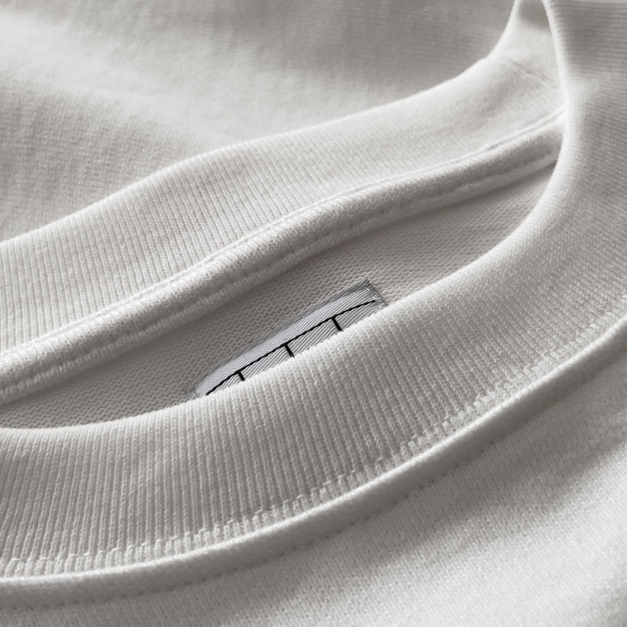 Kırık beyaz (ekru) premium yetişkin t-shirt modelinin yaka ribanasının iç kısmındaki temiz dikiş işçiliğini ve gizli etiket yerleşimini gösteren makro çekim. Kaliteli tok pamuklu kumaş ve iç yaka yapısı, minimalist tasarıma eşlik eden üstün konforu vurgular. Premium duruş ve hassas ciltler için rahatlık sağlayan yapısal detay.
